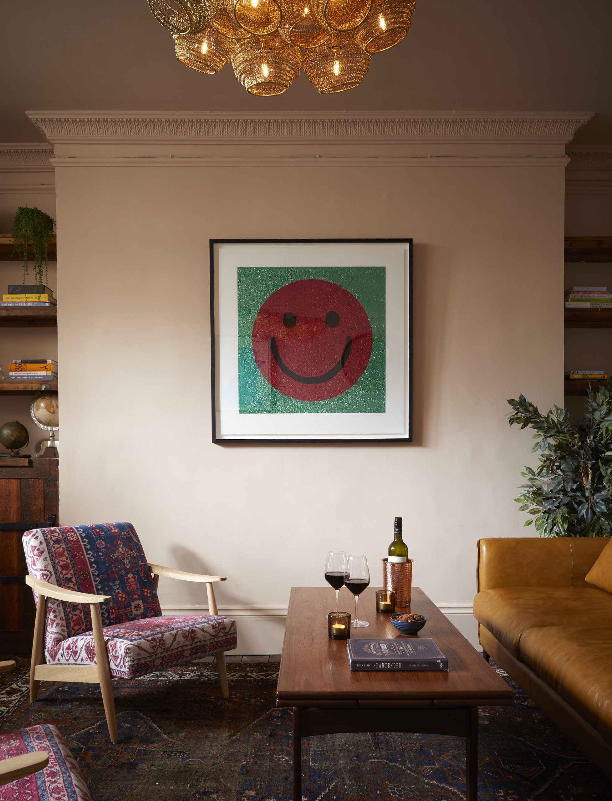 Living room with framed artwork of a smiling red face on green background hanging on wall, armchair with patterned fabric, wooden coffee table with wine bottles and glasses, and green plants.