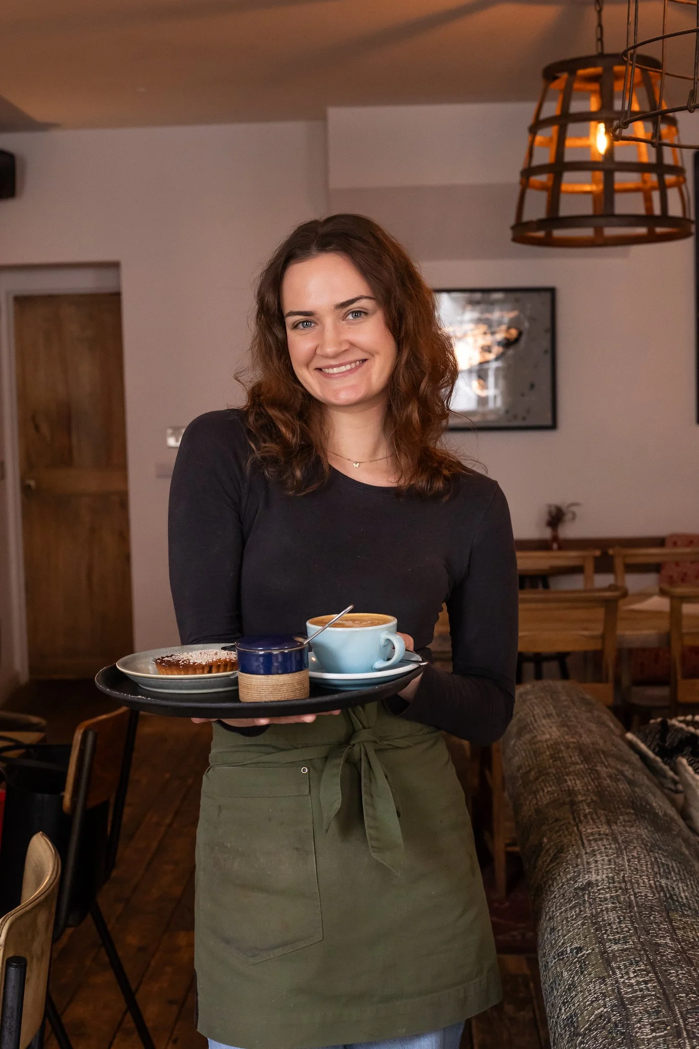 A woman with curly brown hair and a black long-sleeve shirt holding a tray with a mug of coffee or tea, a small blue container, and a plate with a pastry, in a cozy cafe setting.