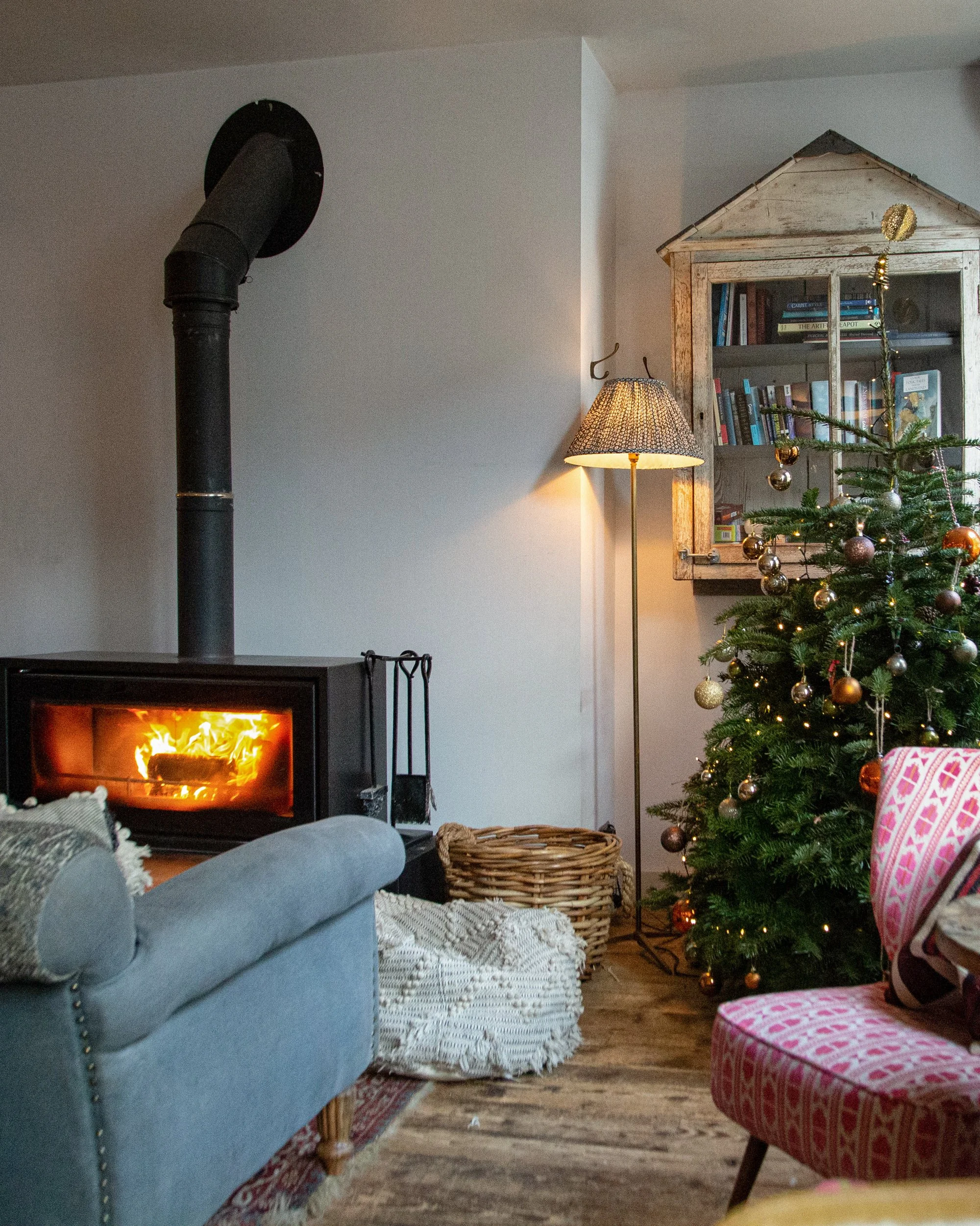Living room with a wood-burning stove, a decorated Christmas tree, a wicker basket, a vintage wooden cabinet with books and decorations, a floor lamp, and patterned furniture.