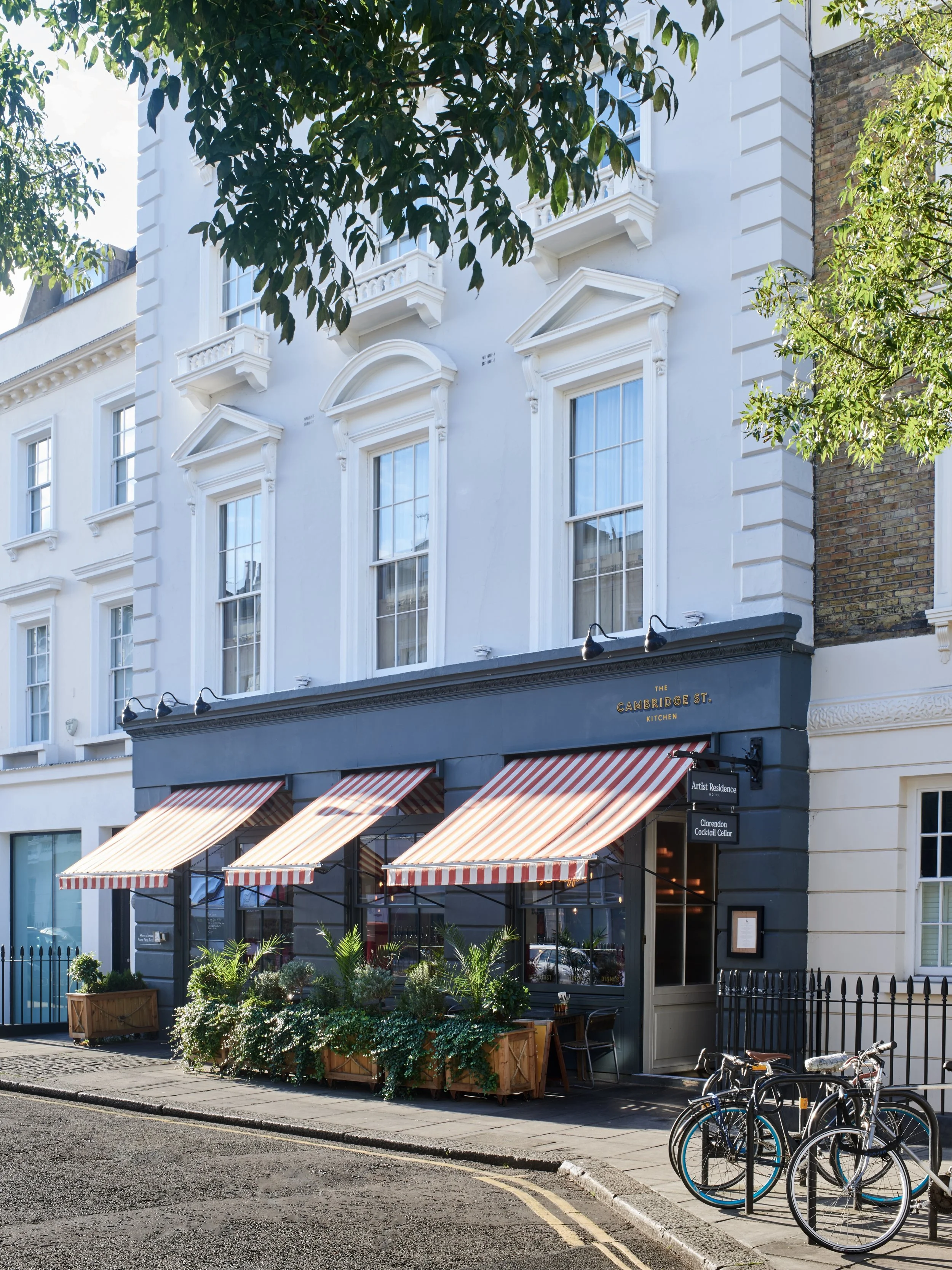 The storefront of a restaurant named 'The Cambridge St. Kitchen' with a black facade, striped red and white awnings, and potted plants outside. There are bikes parked on the sidewalk and trees partially obscuring the upper floors of white residential buildings.