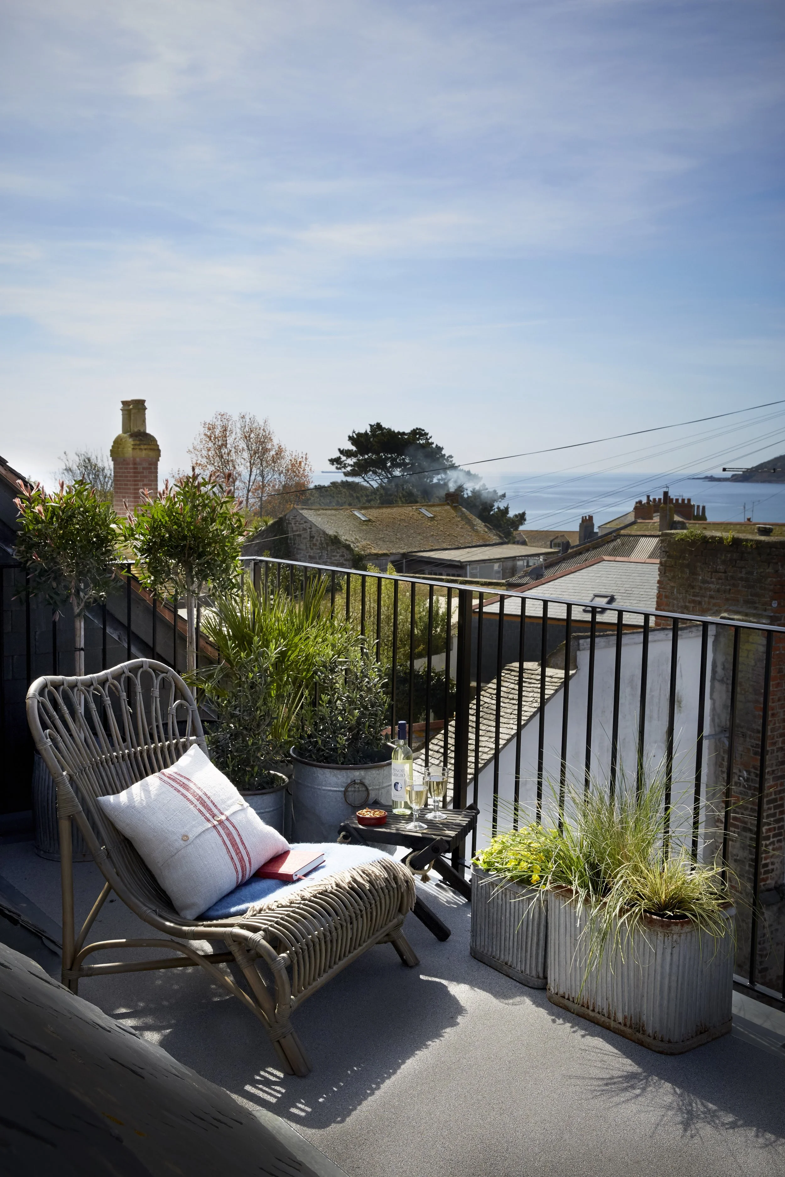 A cozy balcony with a wicker chair, a cushion, and a small table holding a glass of wine, a glass of champagne, and a tart. Potted plants and greenery surround the area, overlooking rooftops, trees, and a distant body of water with smoke or mist in the background under a partly cloudy sky.