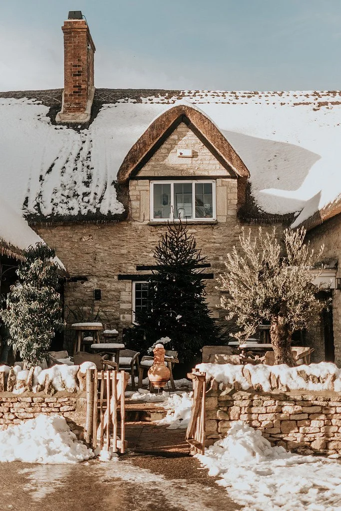 A stone house with snow on the roof and in the yard, featuring a chimney, a Christmas tree, outdoor furniture, and a stone wall with wooden gates.