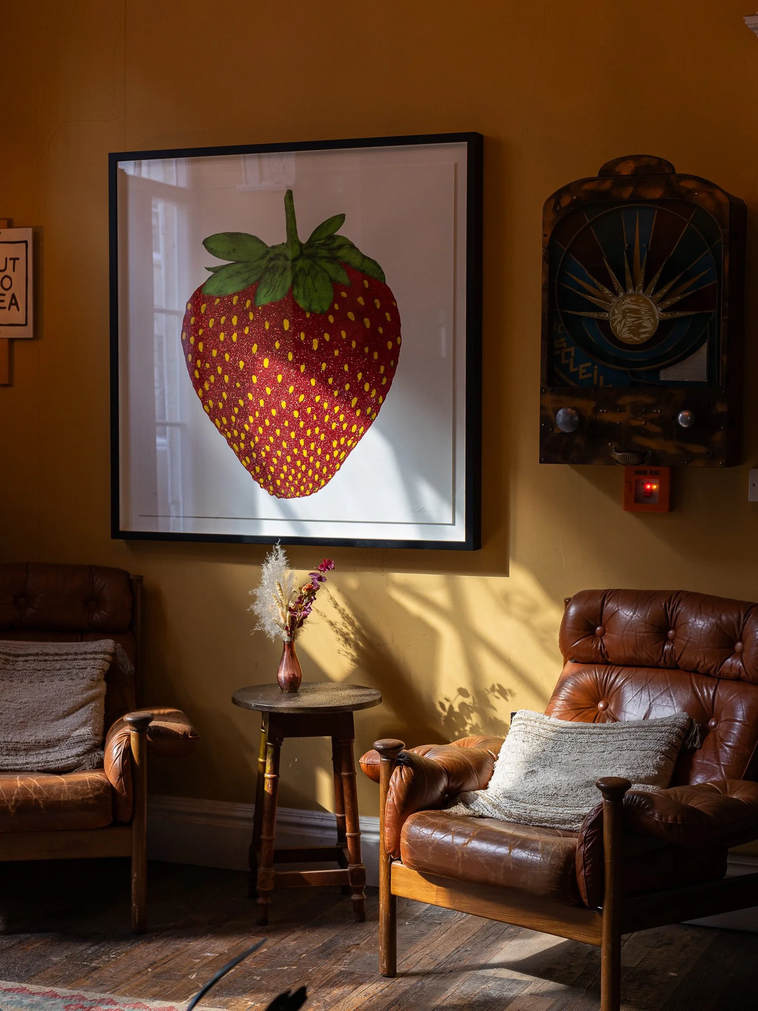 Living room with two brown leather armchairs, one with a grey pillow, a small round wooden table with a pink vase and dried flowers, a framed strawberry artwork on the wall, and a vintage wall clock, all illuminated by sunlight.
