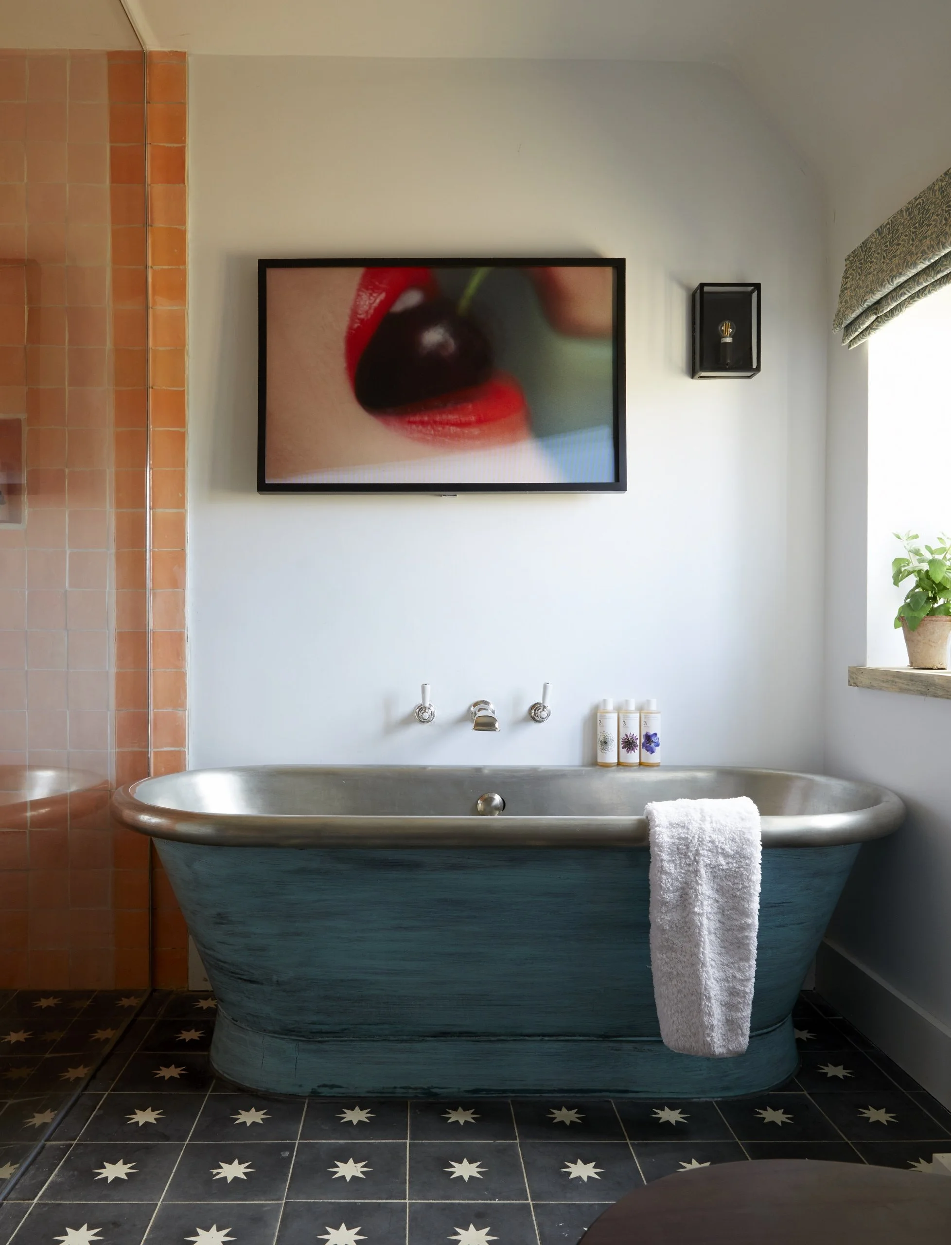 Bathroom with vintage tub, wall-mounted faucets, framed artwork of lips with cherry, small wall shelf with lightbulb, potted plant on window sill, decorated black and white star-patterned floor