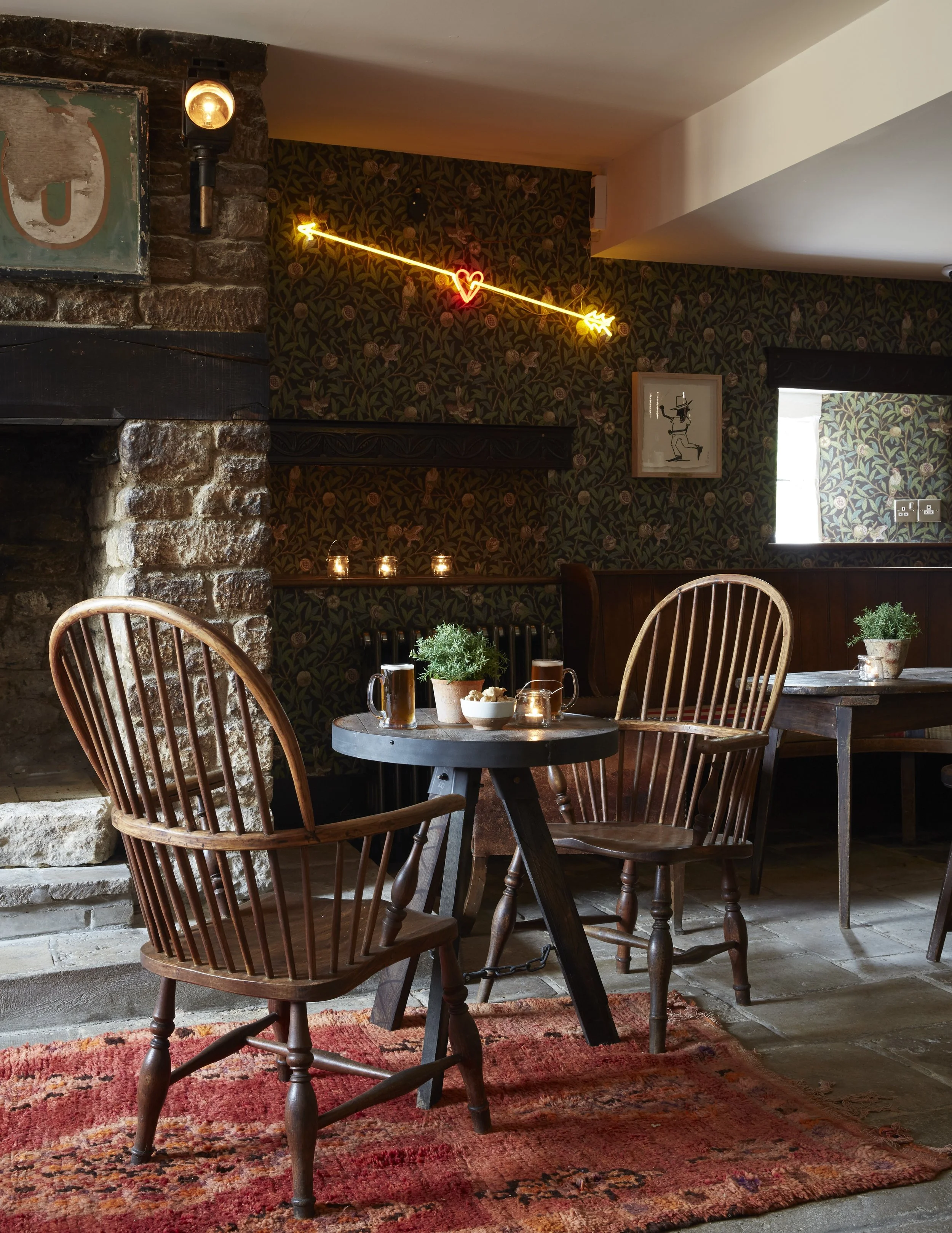 Cozy restaurant interior with wood chairs around a black table holding potted plants, bowls, and drinks. Decor includes a brick fireplace, dim candle lighting, floral wallpaper, and neon wall art with a heart and lightning bolt.