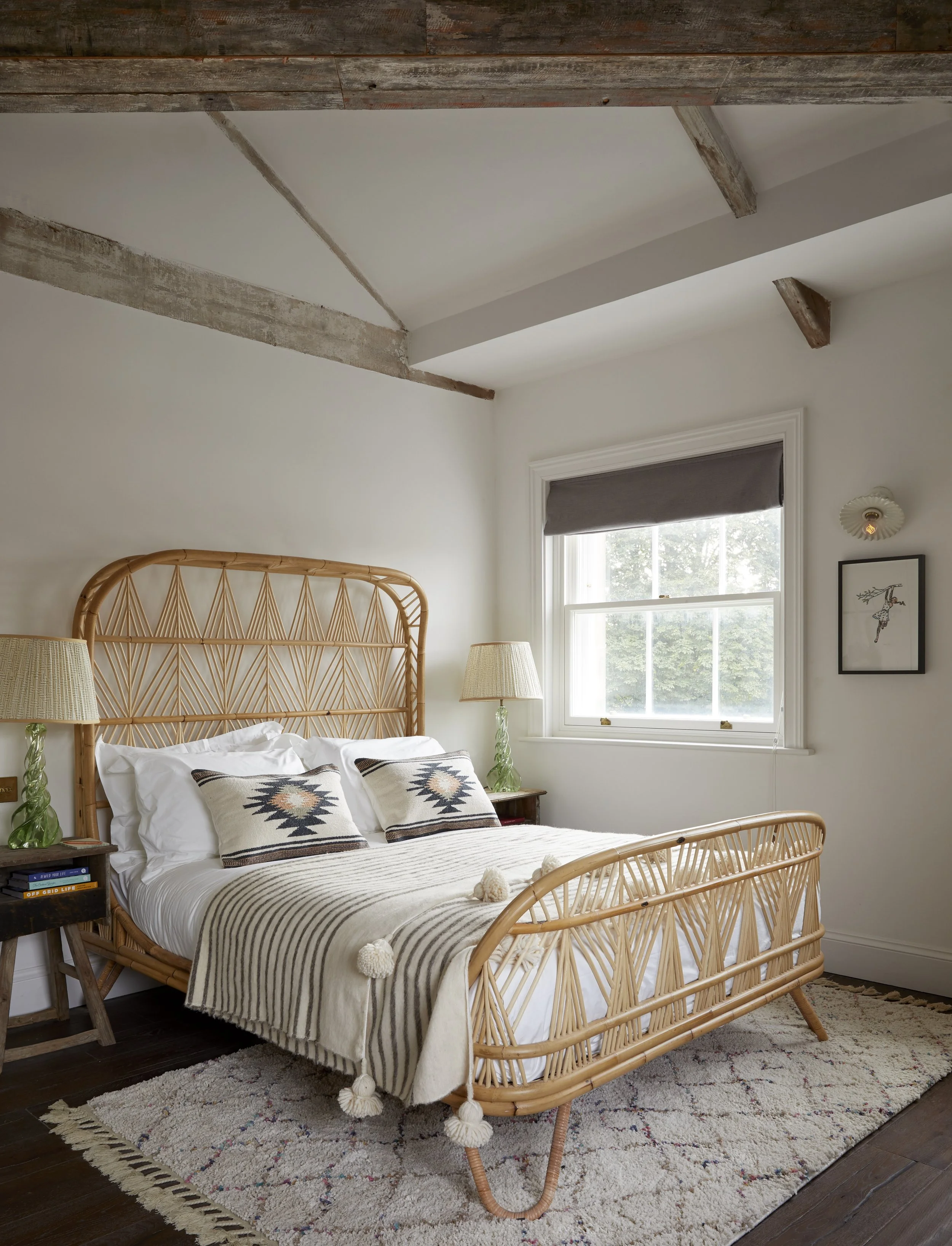A cozy bedroom featuring a wooden bed frame with a woven headboard, decorated with white and patterned pillows. There are matching bedside lamps on each side, and a window with a gray shade allowing natural light. The ceiling has exposed wooden beams and a corner of a framed artwork hangs on the wall. A patterned area rug covers part of the dark wooden floor.
