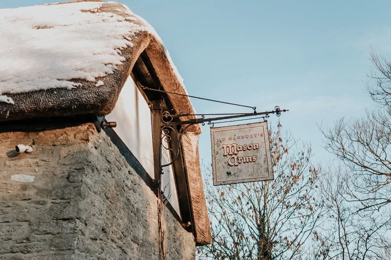 An outdoor sign for 'The Bladensburg Mason Arms' pub hanging from a metal bracket on a stone building, with snow on the roof and leafless trees in the background.