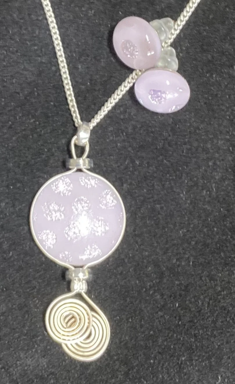 A necklace with a round pendant featuring a light pink background and white floral or lacy design, accompanied by multiple purple and white beads and a spiral-shaped decorative charm at the bottom.