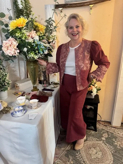 A woman with curly blonde hair, wearing a maroon and gold patterned jacket and matching pants, smiling and standing next to a table with a large floral arrangement, teacups, and decorative items.