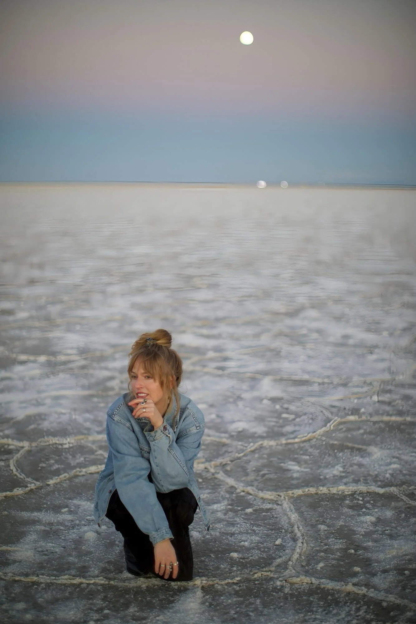A woman with blonde hair in a bun, wearing a denim jacket and black pants, kneeling in shallow ocean water at sunset, with the moon rising in the sky.