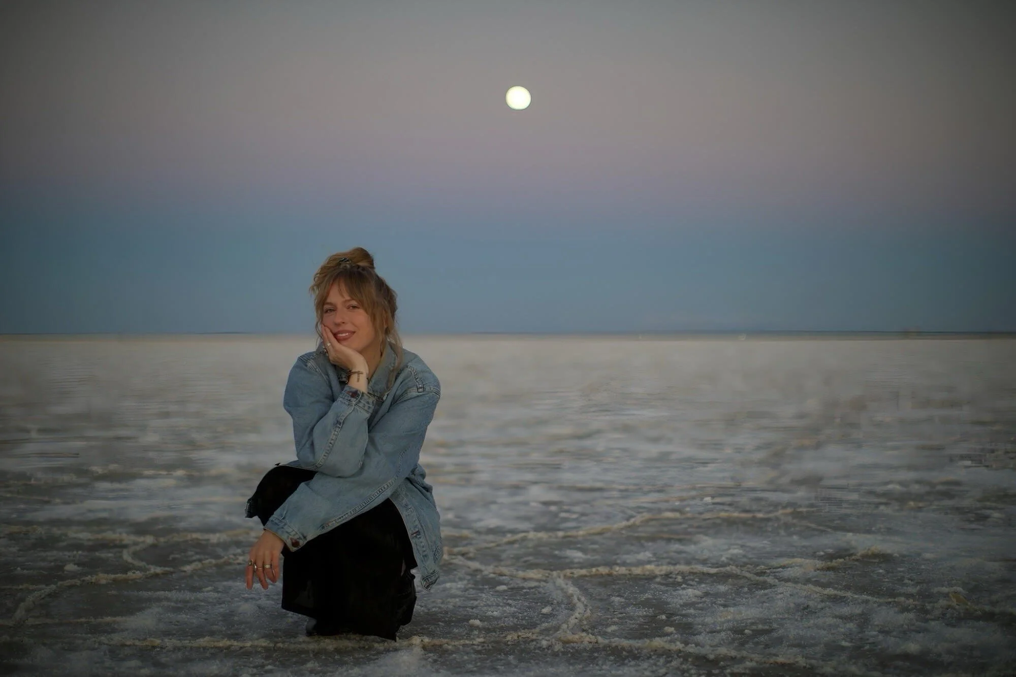 A young woman with blonde hair, wearing a denim jacket and black pants, poses happily on the beach during dusk with the moon visible in the sky and ocean waves in the background.