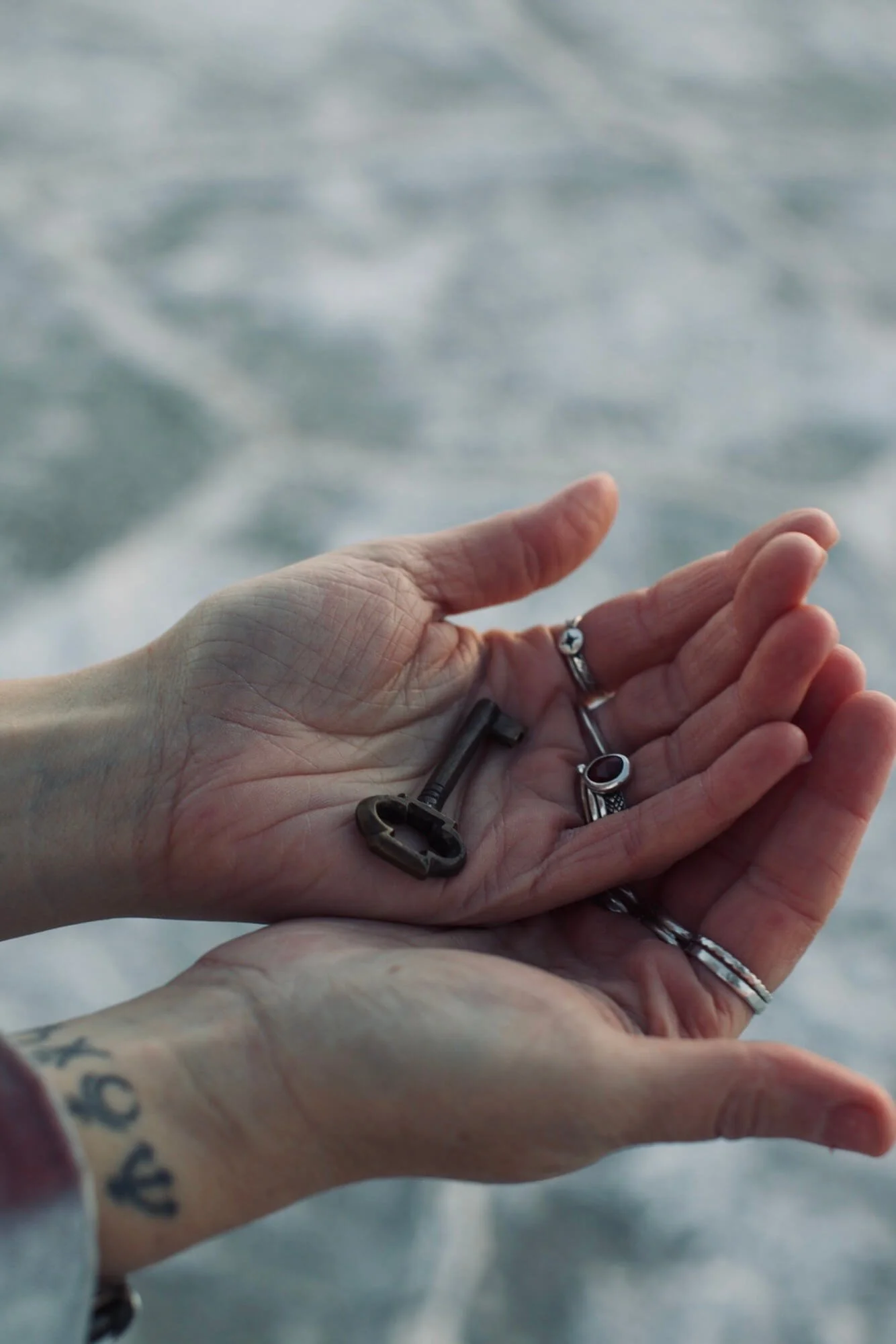 Close-up of a person's hand with rings, holding several old keys, against a blurry background of water.