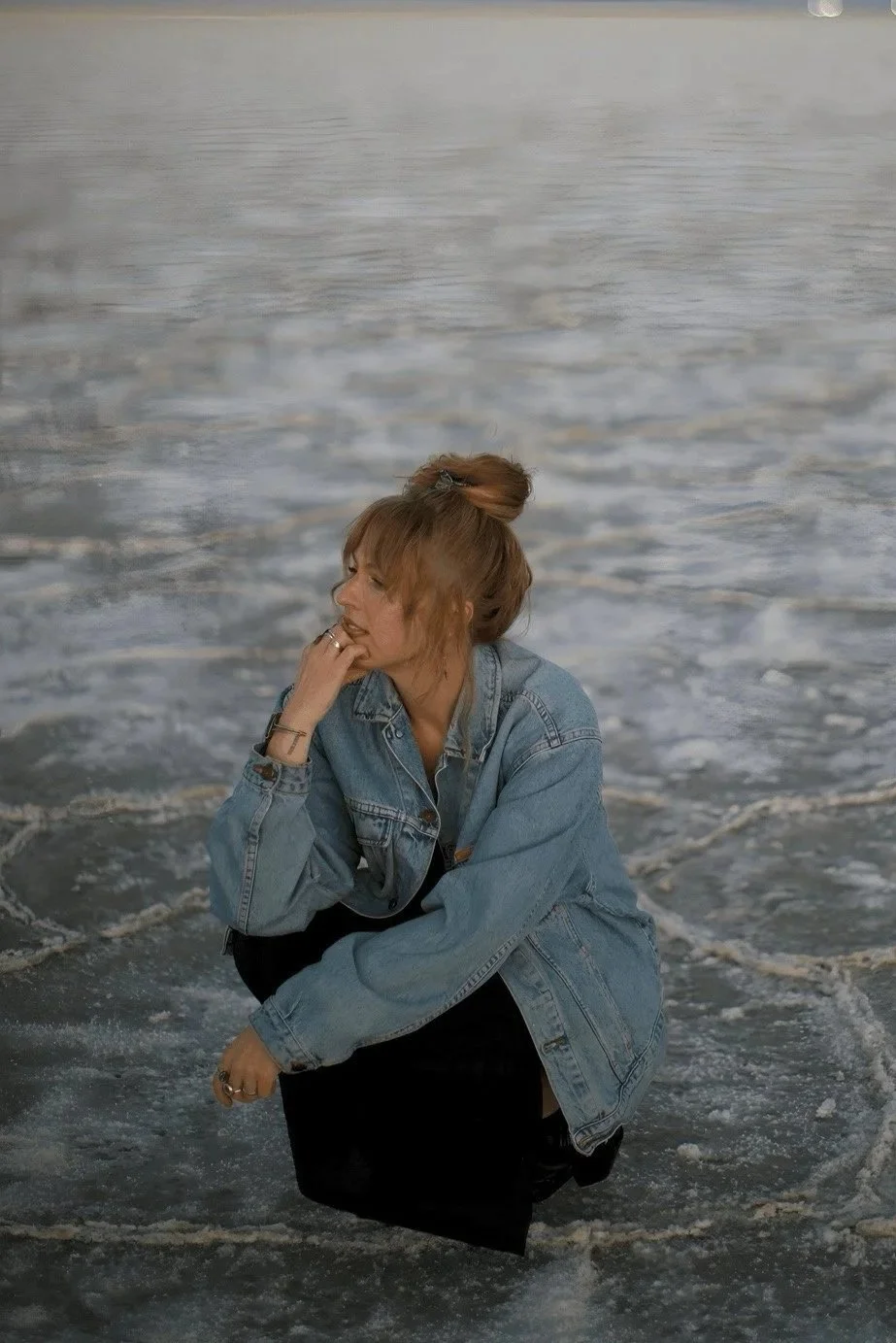 Woman crouching on the beach with ocean waves in the background, wearing a denim jacket and black pants, with her hair in a messy bun, looking to the left.