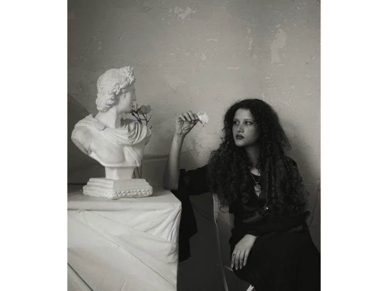 A woman with long curly hair sits next to a bust sculpture on a table, holding a flower in her hand, looking at the sculpture with a thoughtful expression.