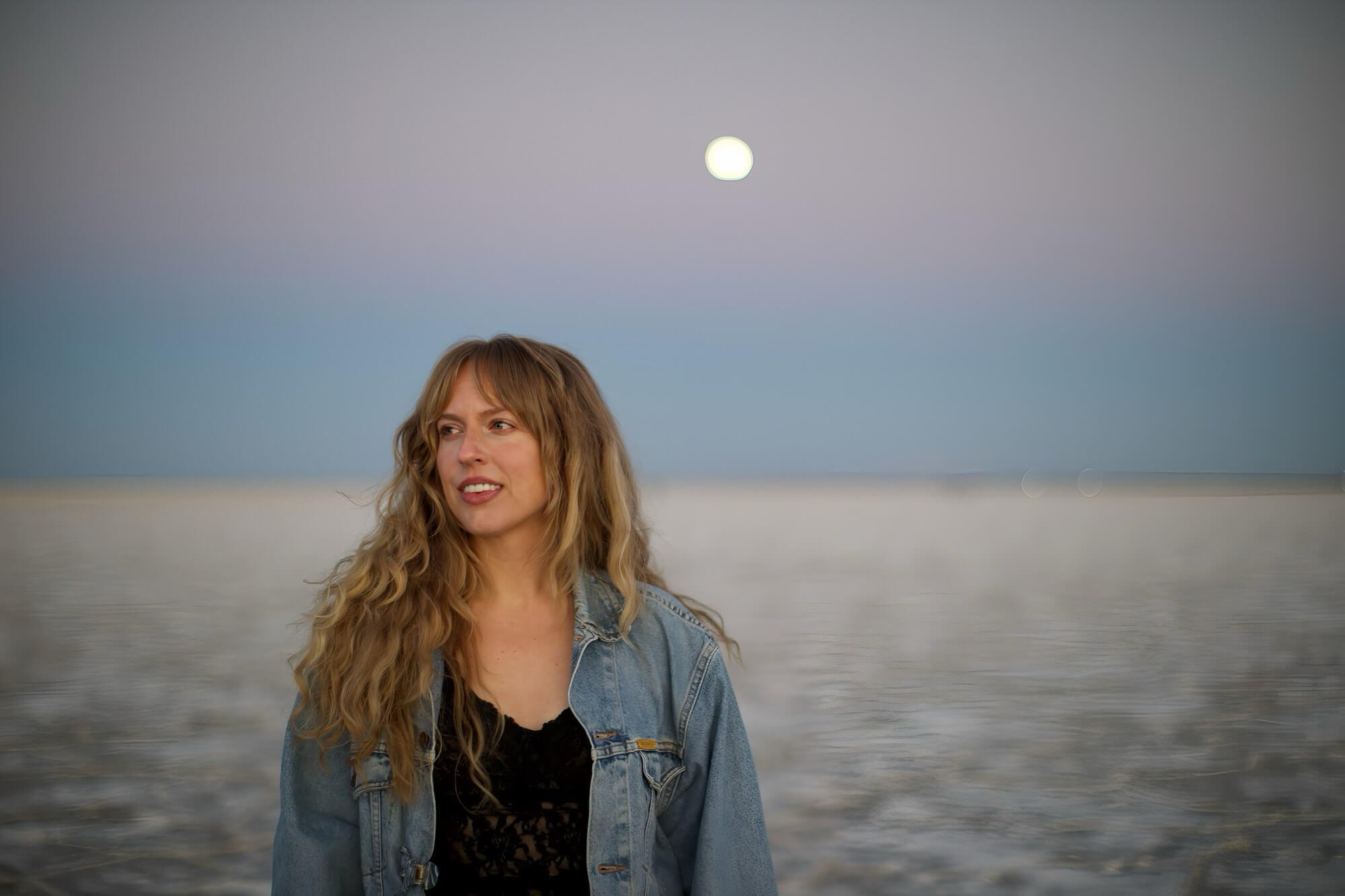 A woman with long curly hair wearing a denim jacket and black top standing near the ocean during sunset or dusk, with the moon visible in the cloudy sky over the water.