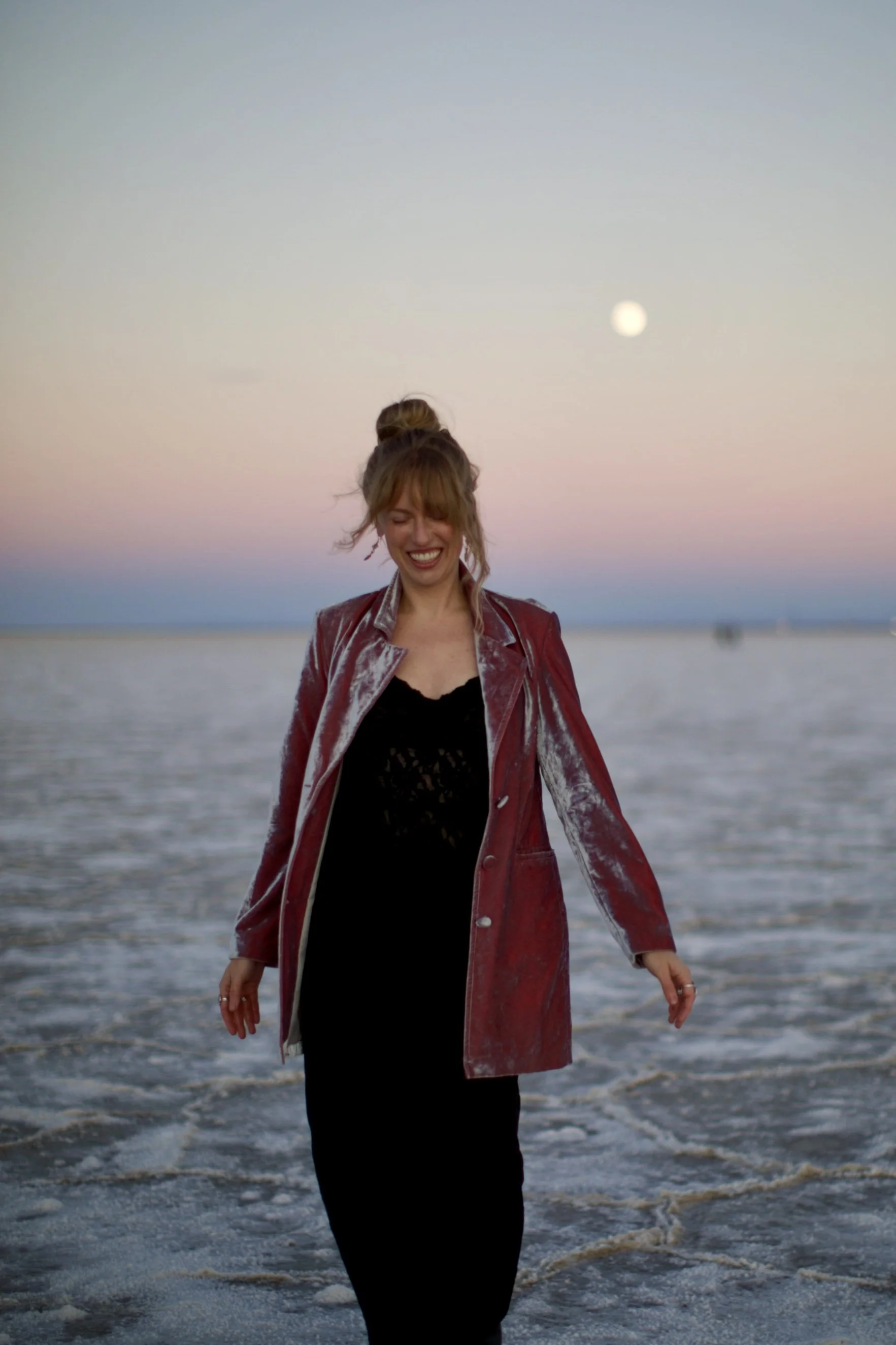 A woman with blonde hair in a messy bun, smiling with eyes closed, wearing a red velvet blazer over a black lace dress, standing on the beach with the ocean waves and a full moon in the sky during sunset or twilight.