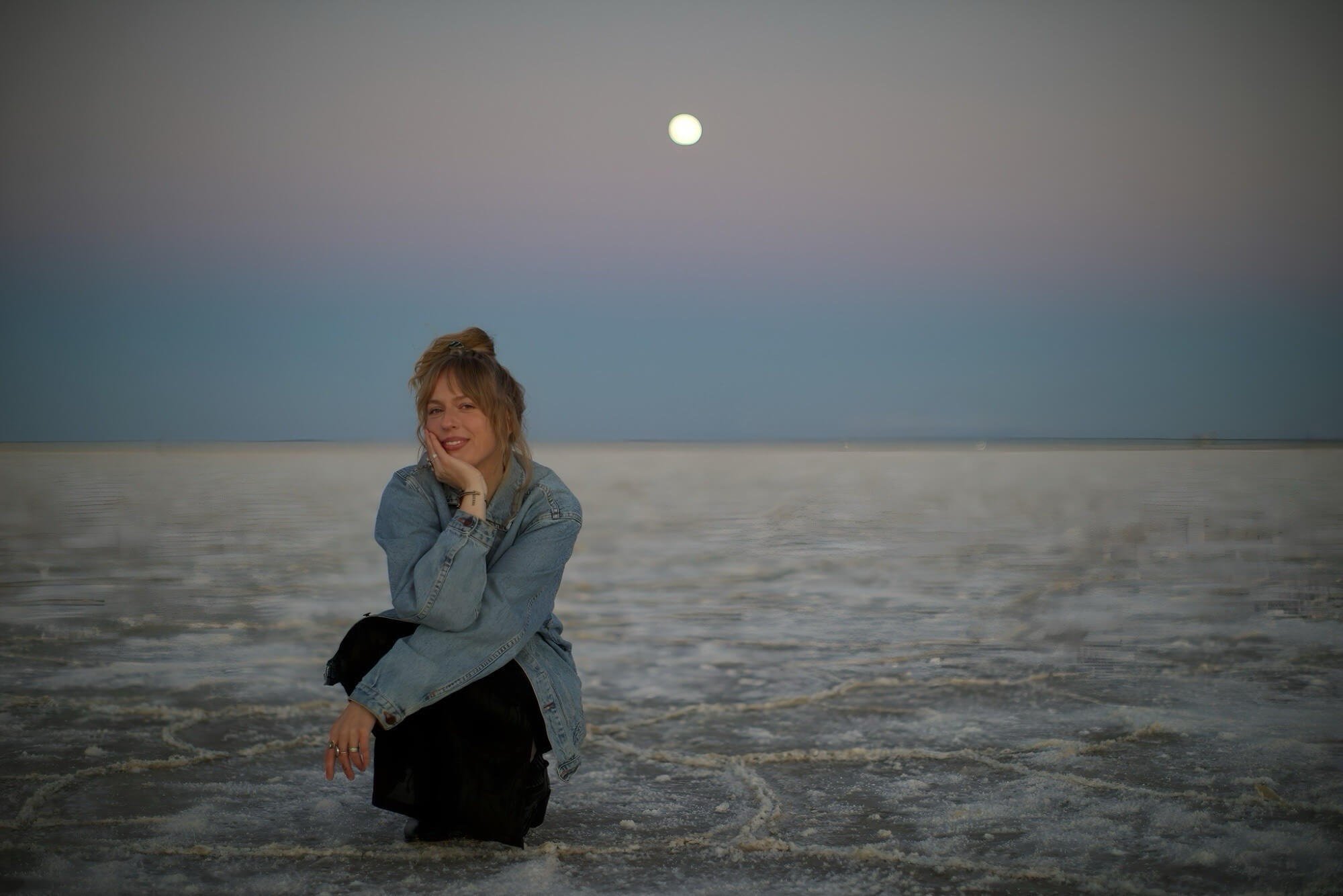 A woman with blonde hair in a bun, wearing a denim jacket and black pants, kneeling in shallow water at the beach during dusk, with a full moon in the sky above.