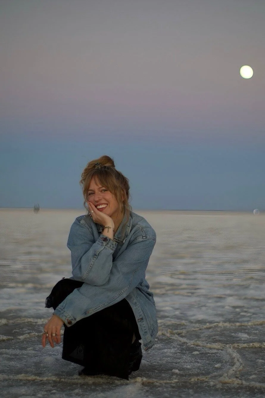 A young woman with light brown hair in a bun, smiling and crouching in shallow water at the beach during dusk, with a full moon in the sky.