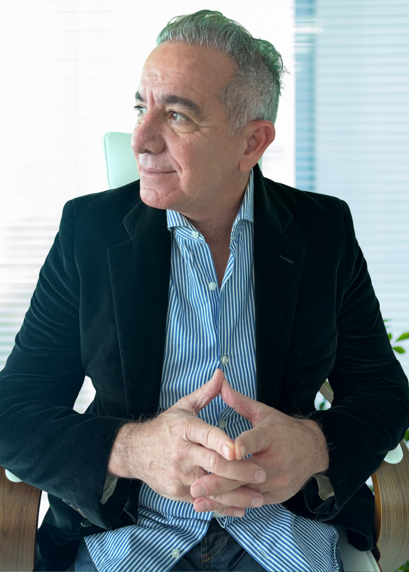 A middle-aged man with gray hair, wearing a dark blazer and a blue and white striped shirt, sitting at a table with his hands clasped together, looking to the left, in a bright office setting.
