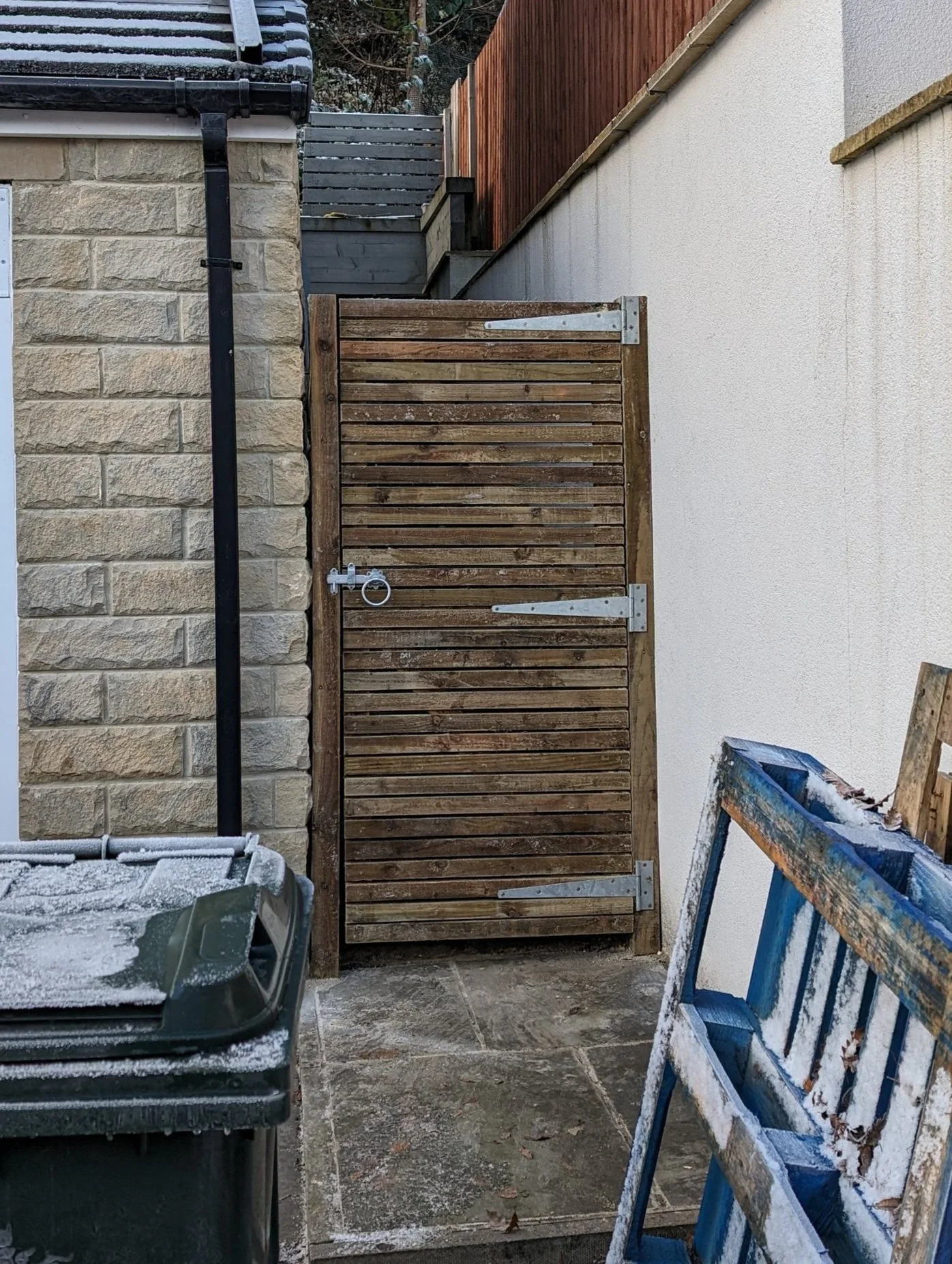 Wooden gate with metal hinges and latch in an alleyway, surrounded by a stone wall, a green garbage bin, and a blue pallet, with a dusting of snow on the surfaces.