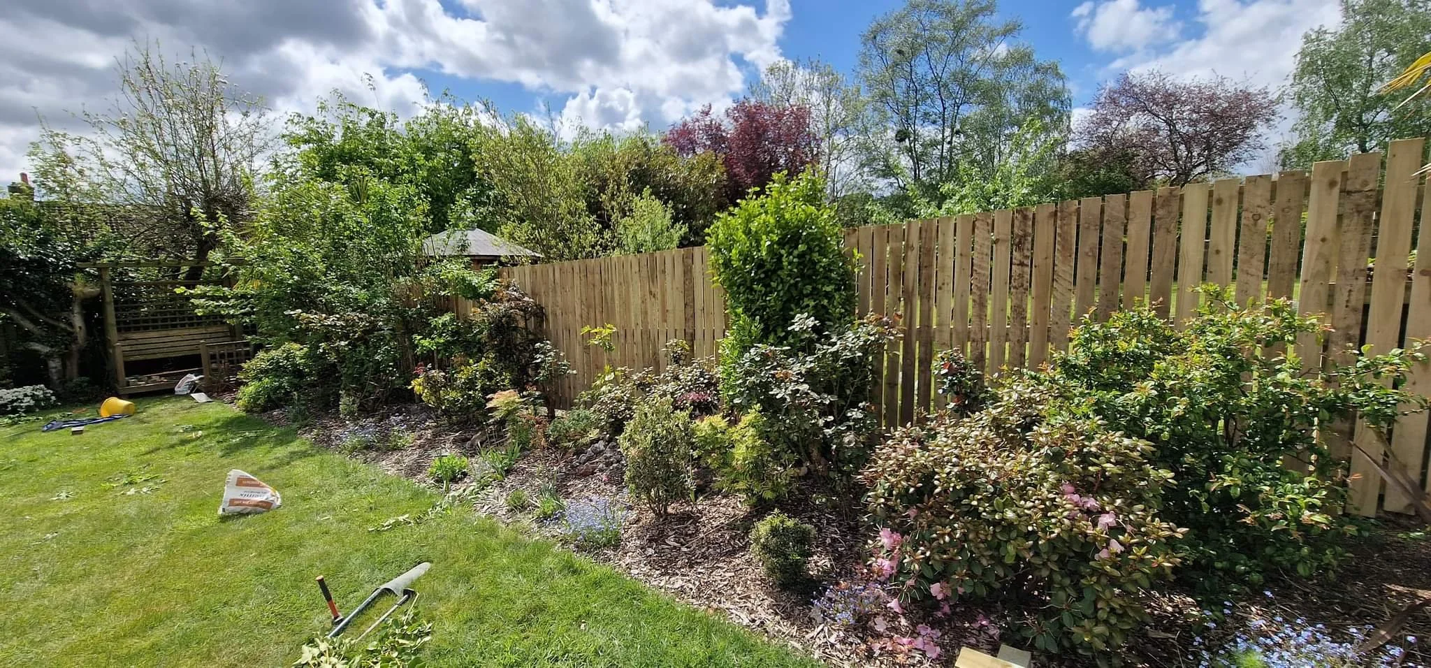 Backyard garden with wooden fence, various plants and shrubs, a wooden pergola, tools and materials on the grass, cloudy sky.