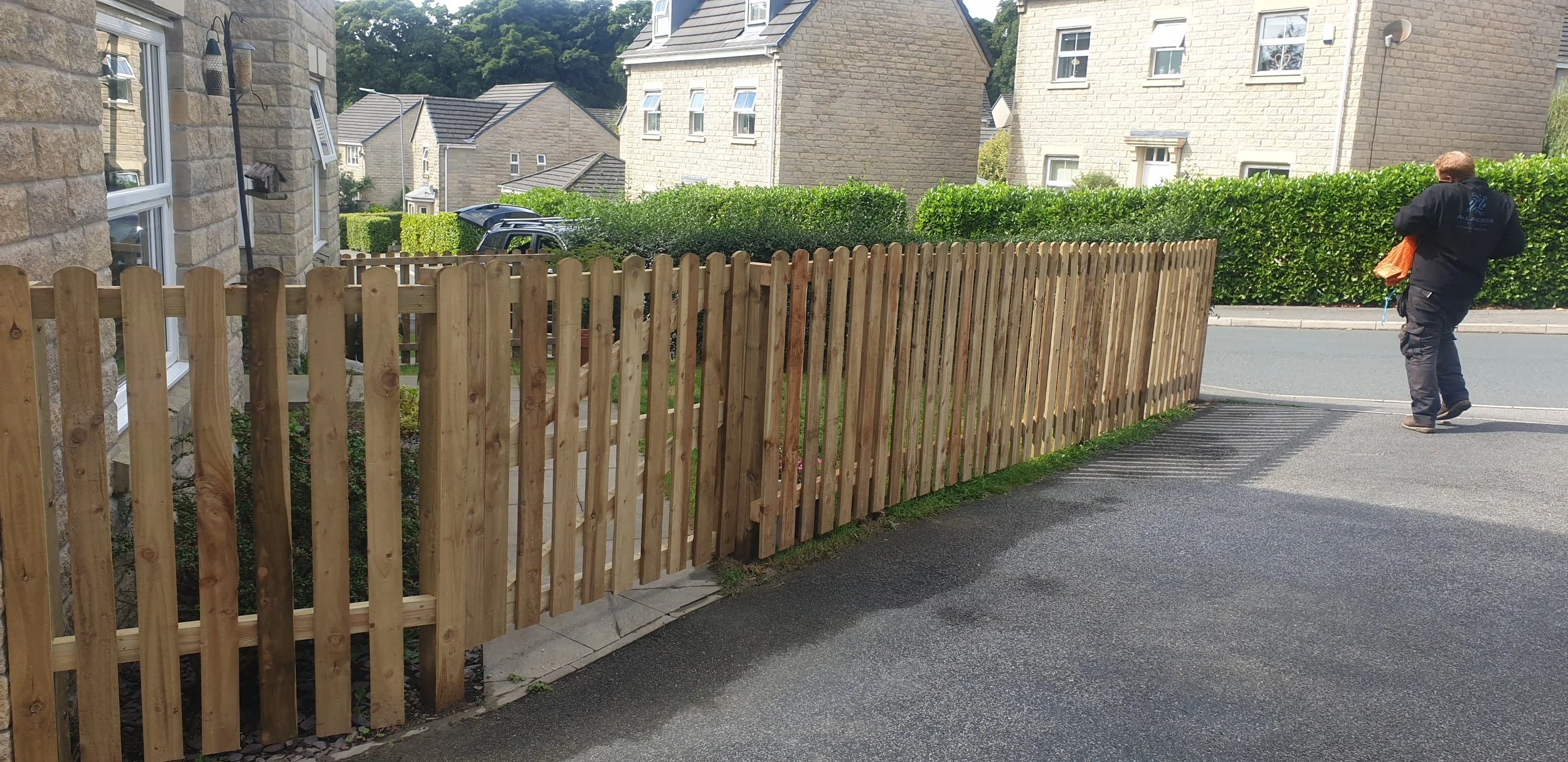 Wooden picket fence in front of stone houses on a residential street, with a person walking away.