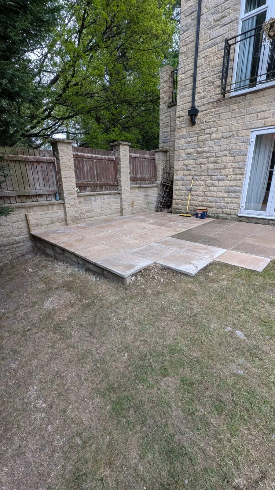 A newly installed backyard patio with beige and tan stone tiles next to a brick house, bordered by a wooden fence and surrounded by trees.