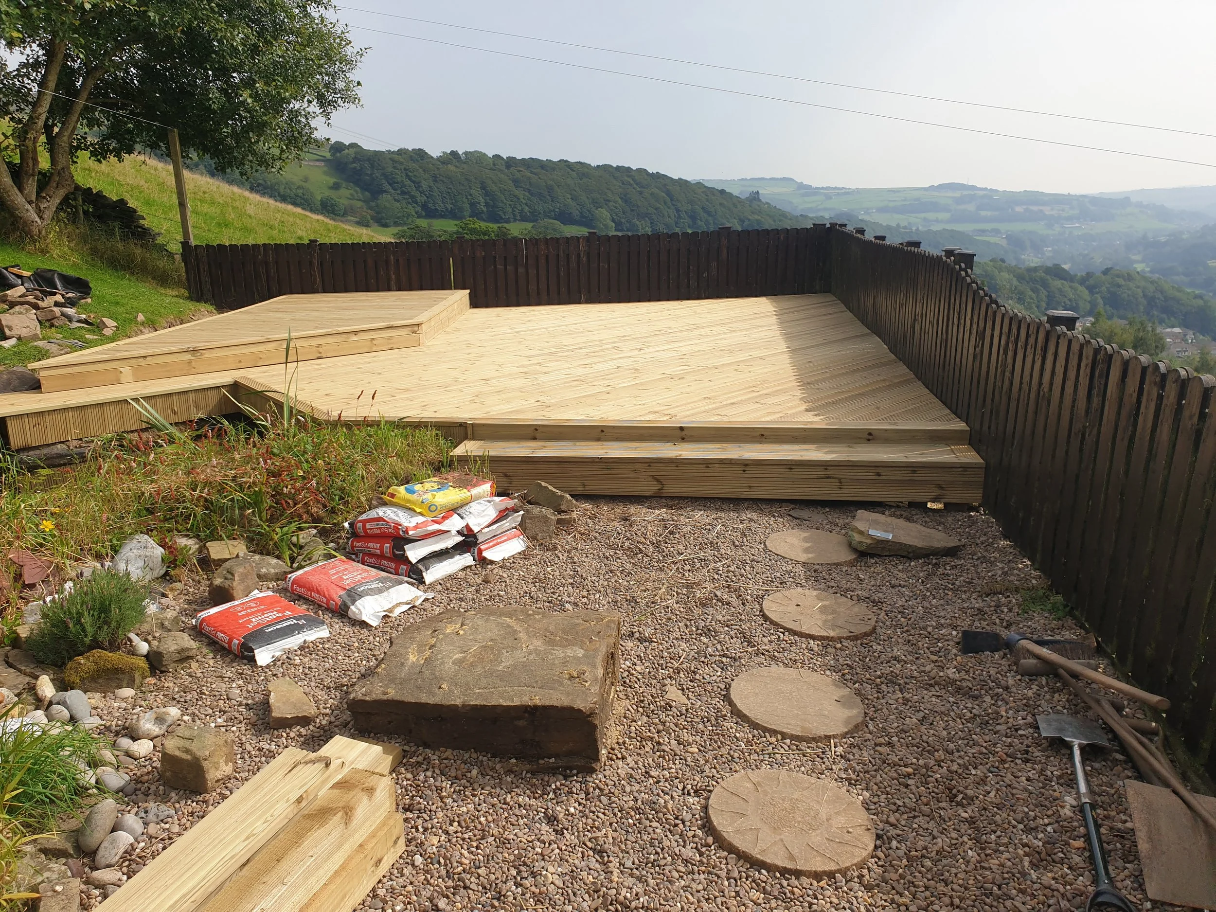 Wooden deck with surrounding fence, bags of construction materials, flowerbed, stone pathway, and garden tools, located in a scenic countryside setting.