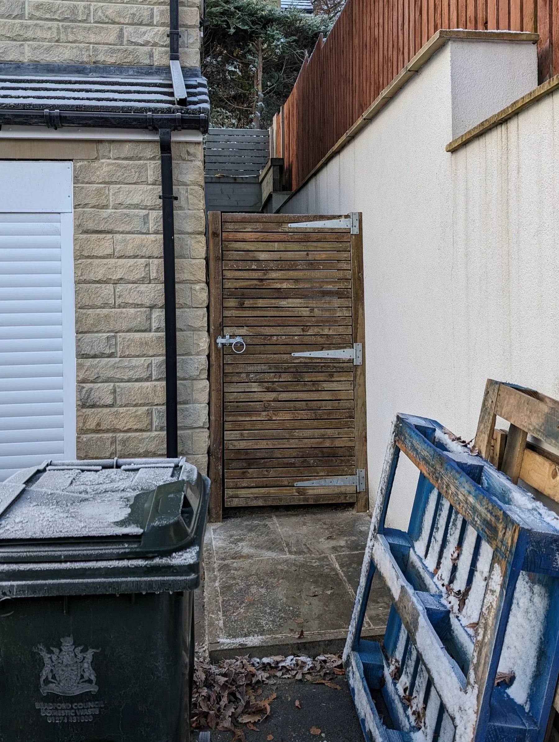 Wooden gate in narrow alley with stone wall on one side and white plaster wall on the other, adjacent to a frosted trash bin and blue pallet.