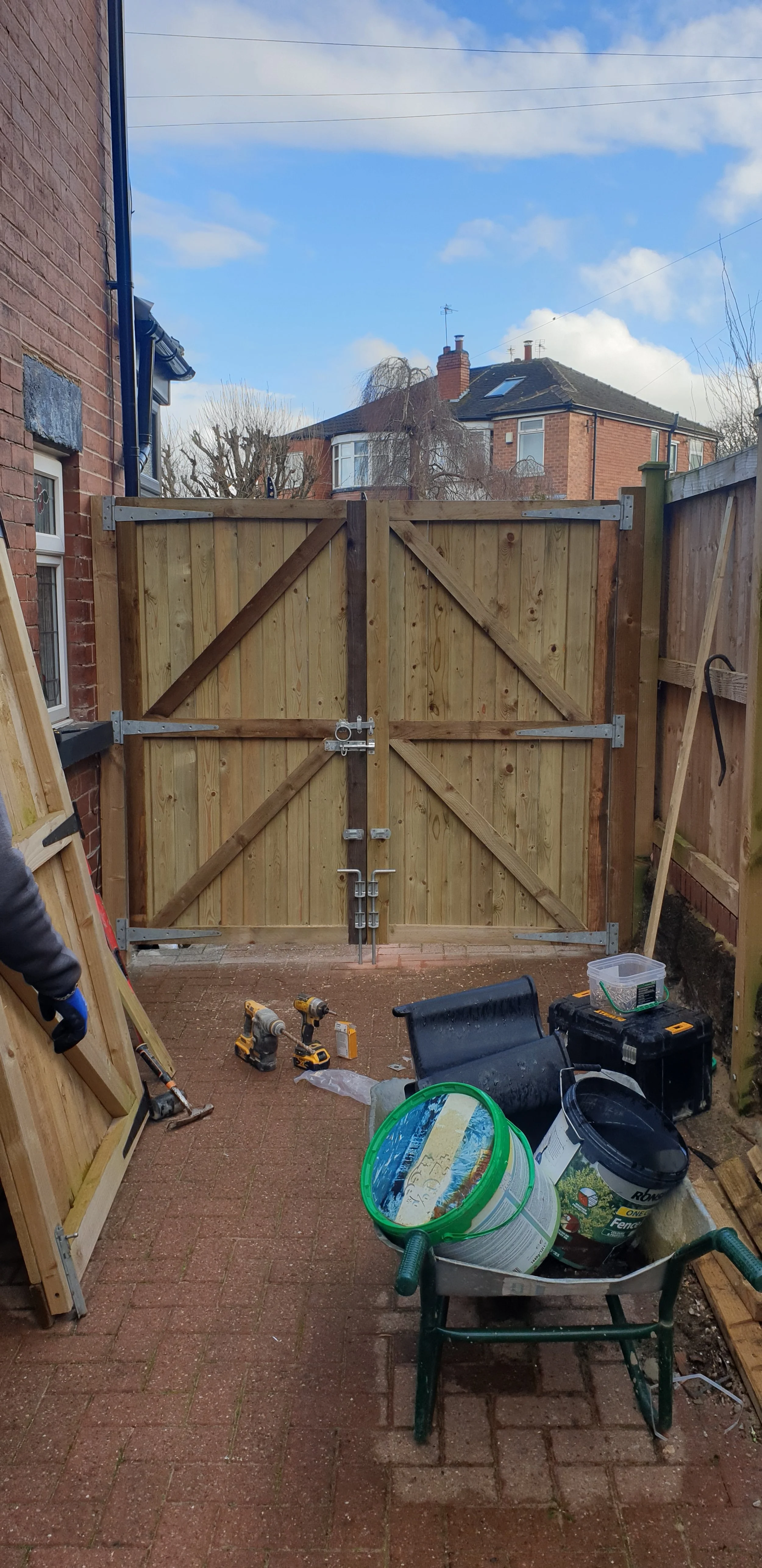 Wooden double gate with metal hinges and lock, surrounded by brick walls and a fence. Near the gate, tools like drills, buckets, and a wheelbarrow are scattered on a brick patio. Two-story brick house visible in the background under a blue sky with clouds.