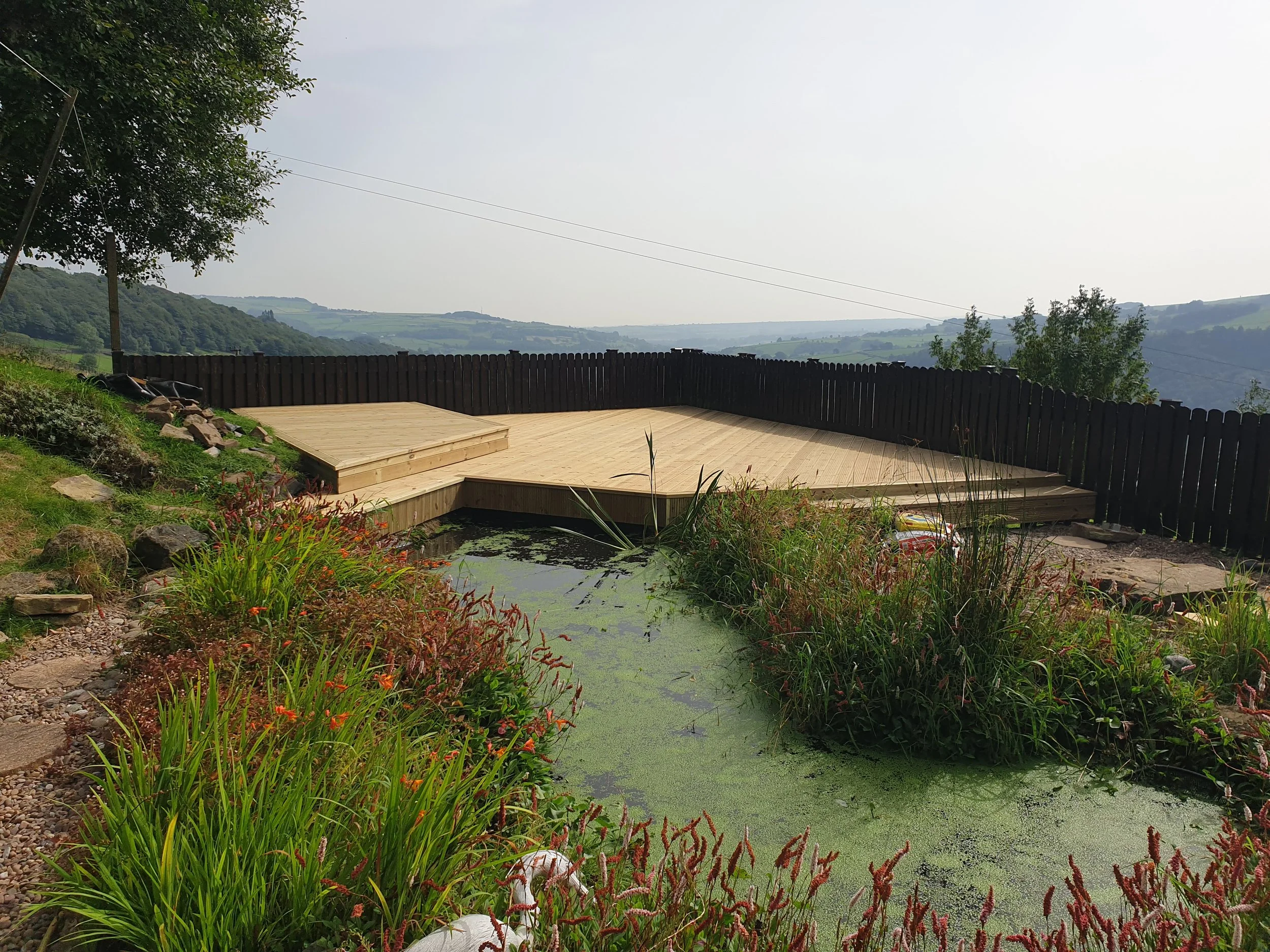 Wooden deck with fence overlooking scenic landscape and pond with plants