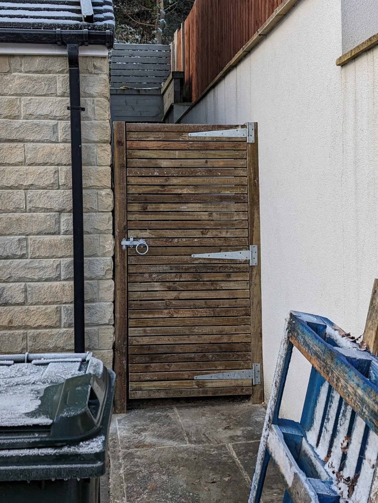 Wooden gate with metal hinges, adjacent to a stone building and a white wall. Nearby is a green outdoor trash bin and a blue wooden crate, both with frost.