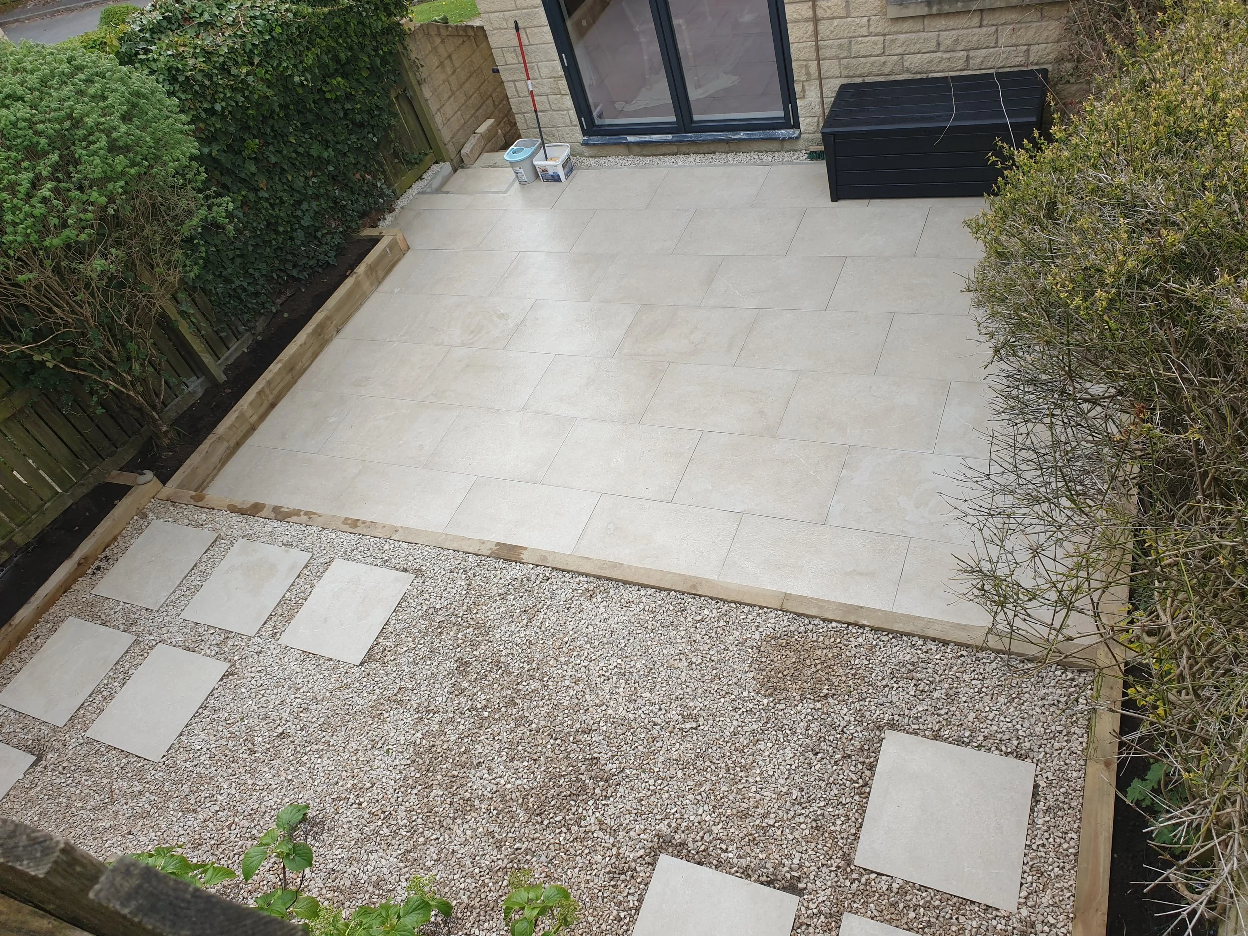 Outdoor patio with large light-colored tiles, gravel pathway with stepping stones, black storage box, garden hedges, and plants.