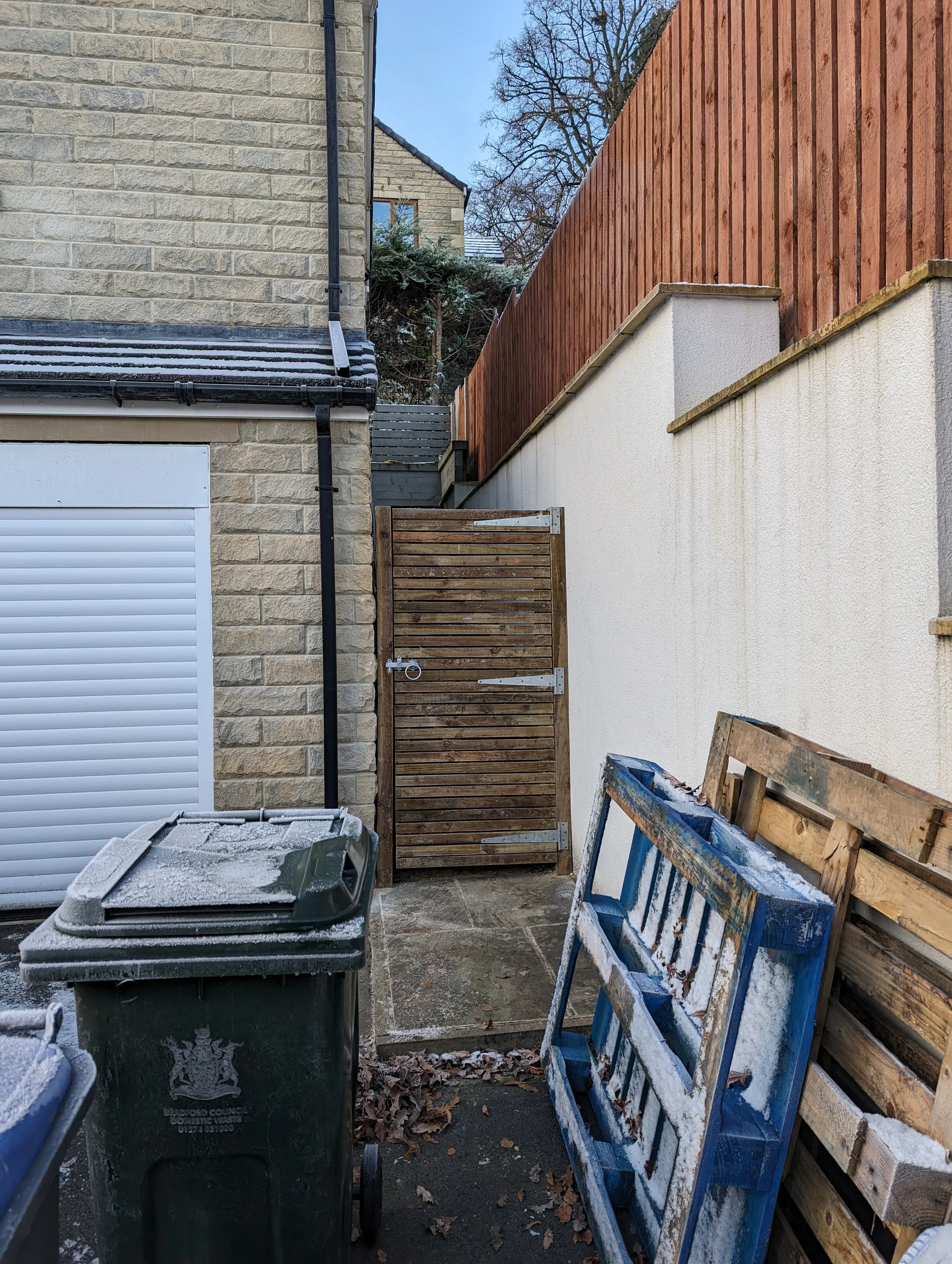 Outdoor area with a wooden gate, a green garbage bin, wooden pallets leaning against a wall, a brick building, and a wooden fence.