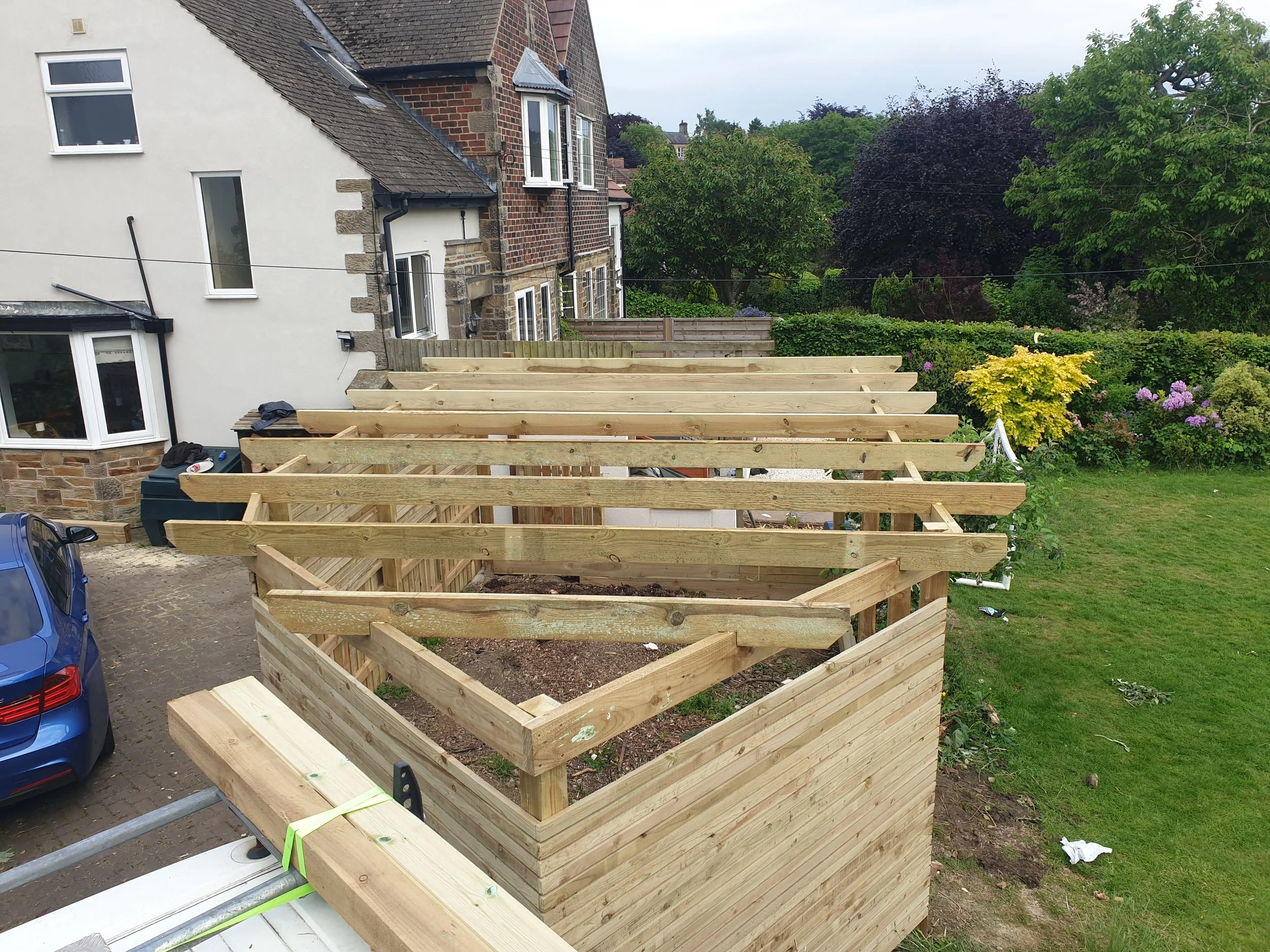 Construction of wooden garden structure near a house with a blue car parked nearby and a garden with trees and shrubs in the background.