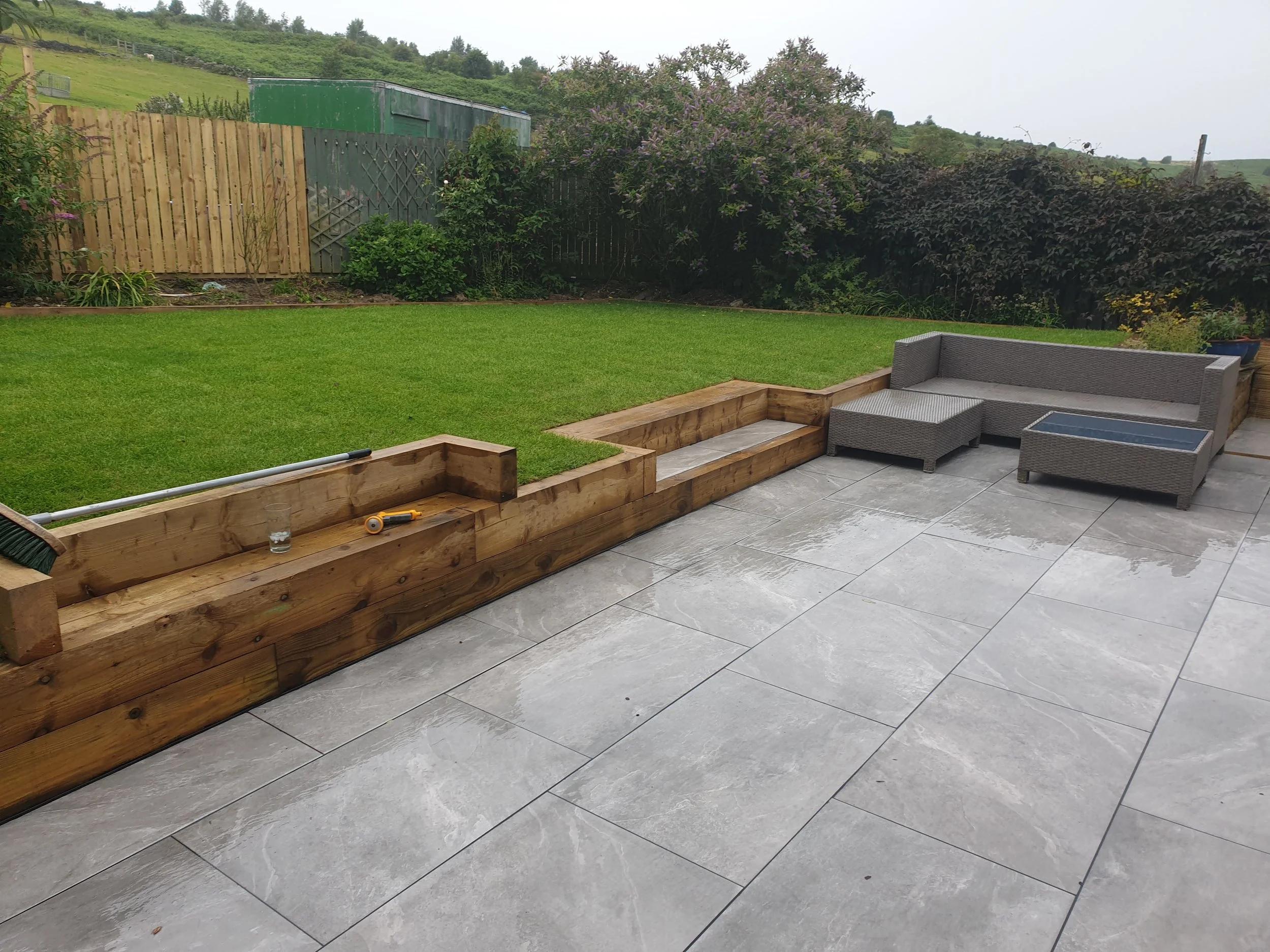 Outdoor patio with gray tiles, wooden planters, and a woven sofa set with an ottoman and coffee table, overlooking a lawn with fenced backyard.