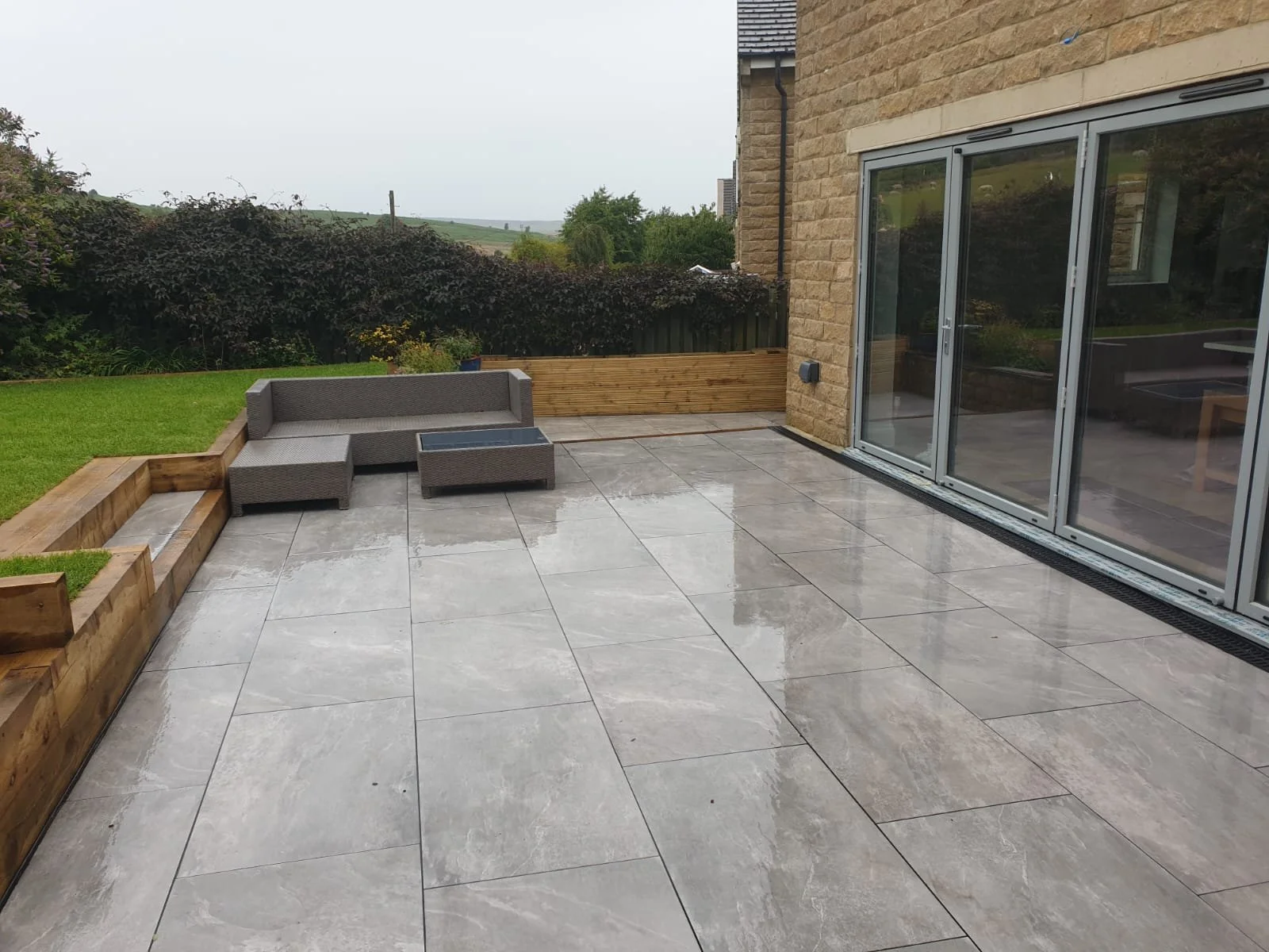 Empty patio with wet tiles, gray outdoor sofa, and a small table, with a small grassy yard, bushy trees, and a countryside landscape in the background.