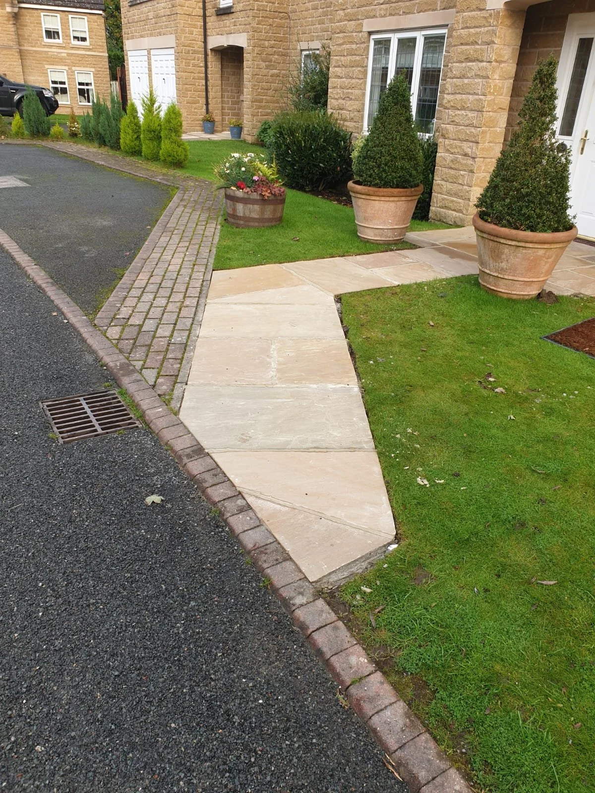 Front entrance of a house with a paved walkway, potted plants, a grassy lawn, and parked cars in the background.
