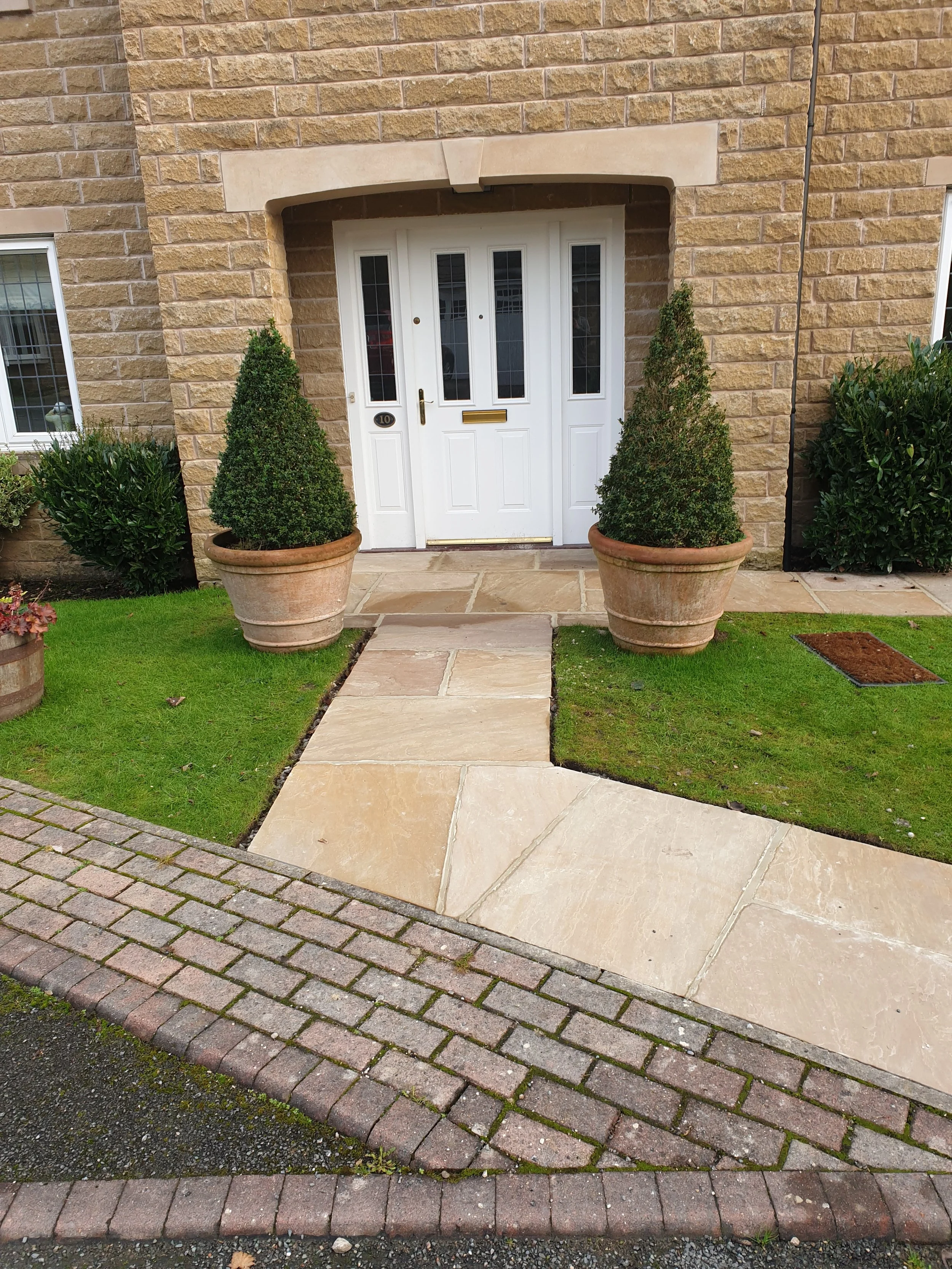 A house entrance with a white door, flanked by two potted topiary trees. The entrance has a stone walkway and brick border, leading to a grassy area.