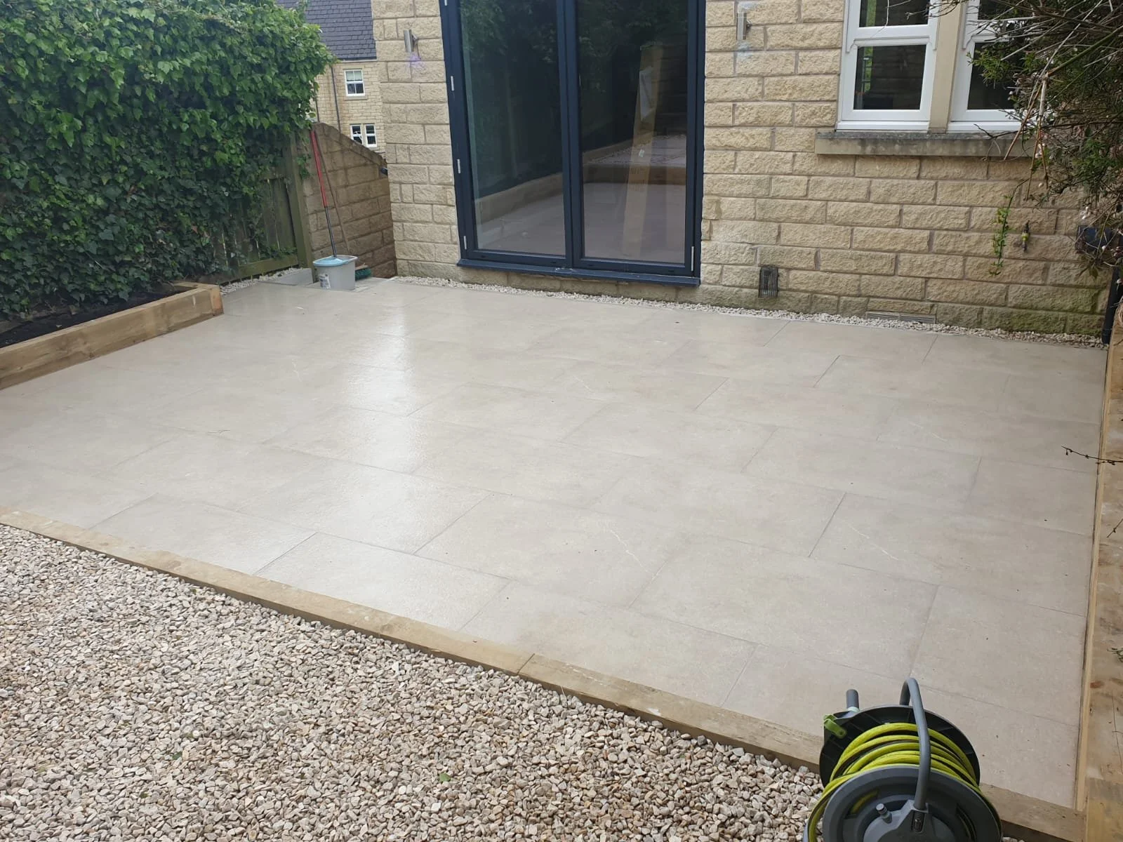 Recently installed beige paving slabs on a backyard patio, bordered by gravel and garden bed with wooden edging. A sliding glass door into the house and a small window in the brick wall are visible. Gardening tools and hose reel are in the scene.