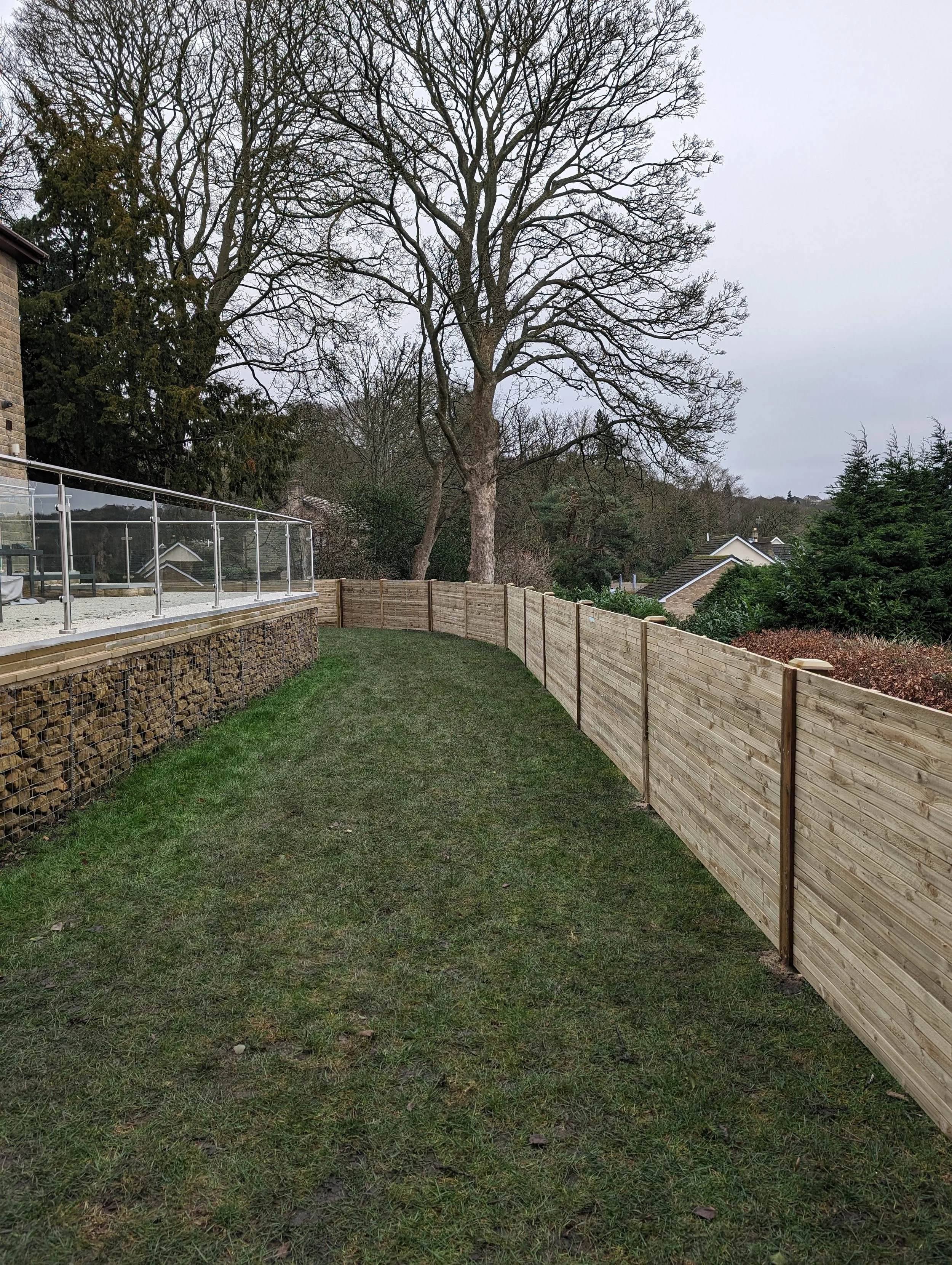 Backyard with a wooden fence, dry grass, and large bare trees; stone wall and glass railing on the left side.