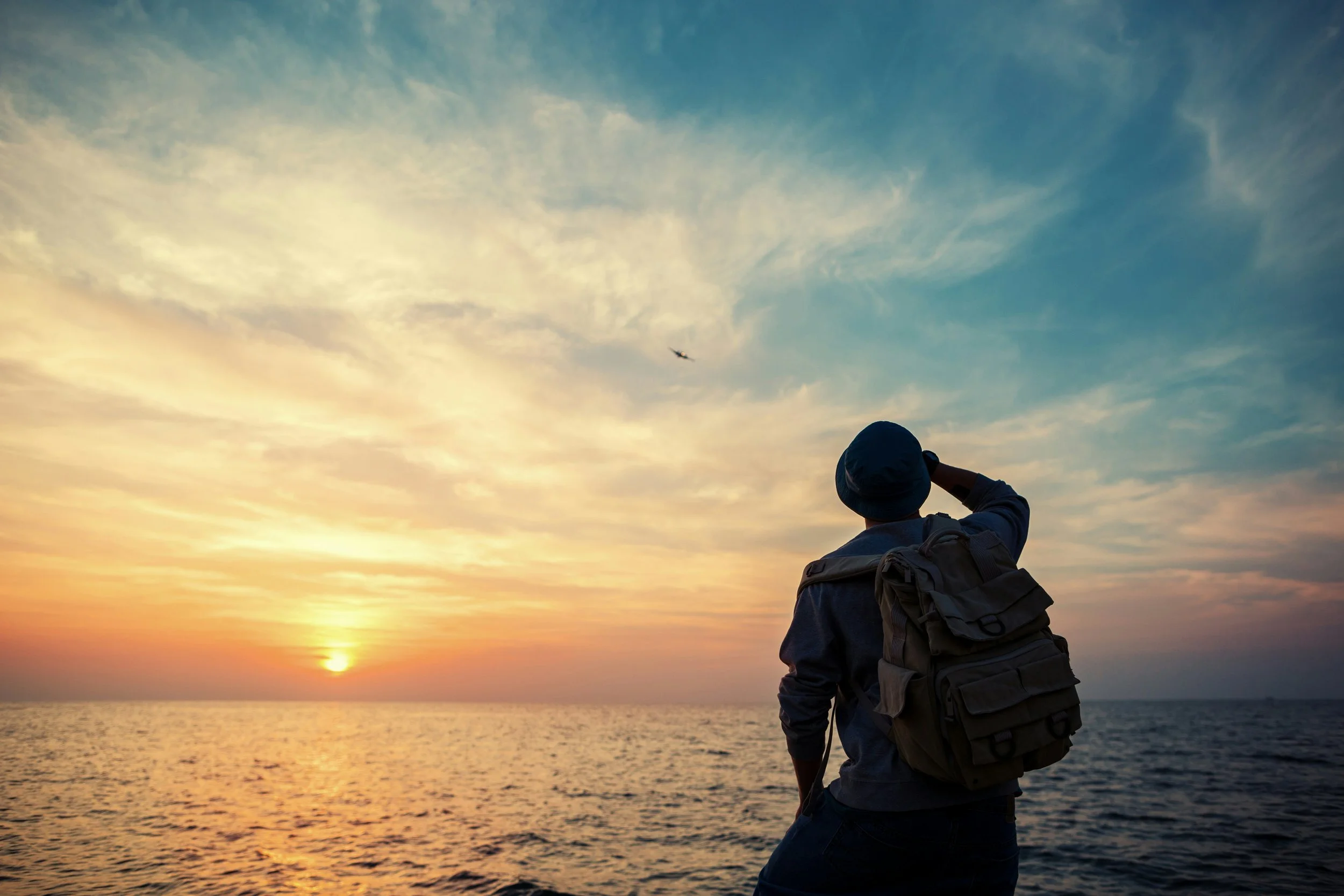 Traveler with a backpack looking out over the ocean at sunset as an airplane flies overhead.