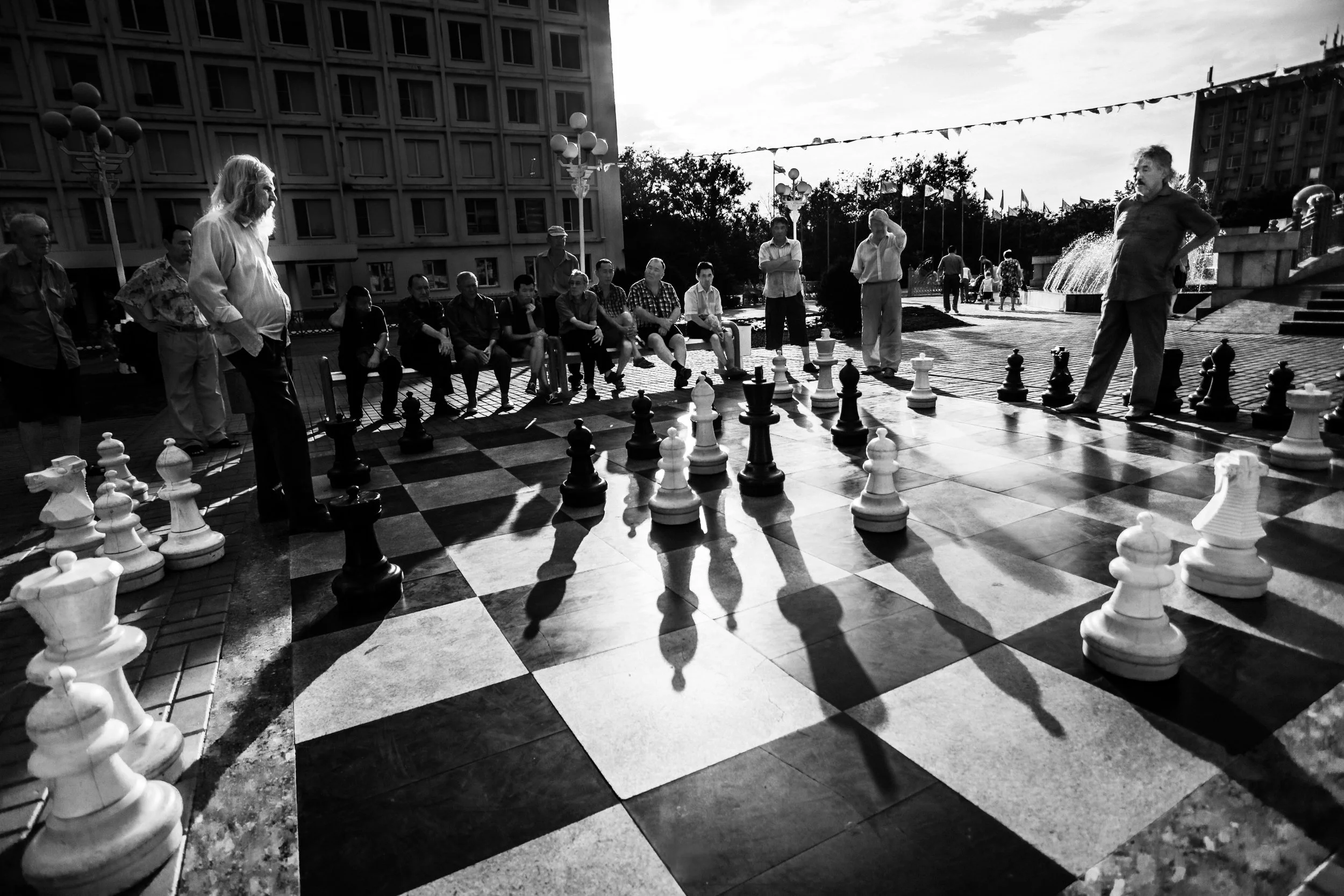 Two players and spectators around a giant outdoor chessboard in black and white, representing strategic struggle and cultural influence.