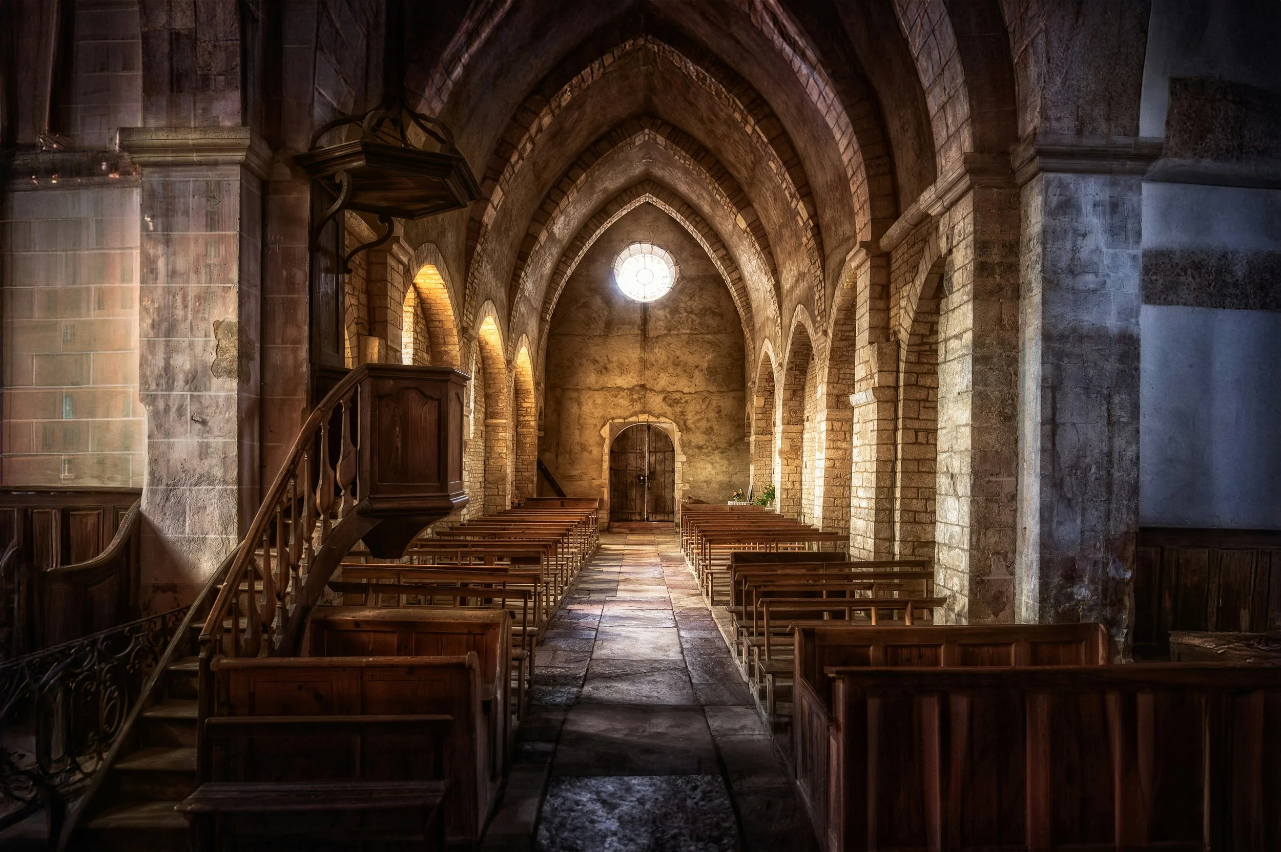 Sunlit interior of a medieval stone church featuring gothic arches, wooden pews, a raised wooden pulpit, and a central stone aisle.