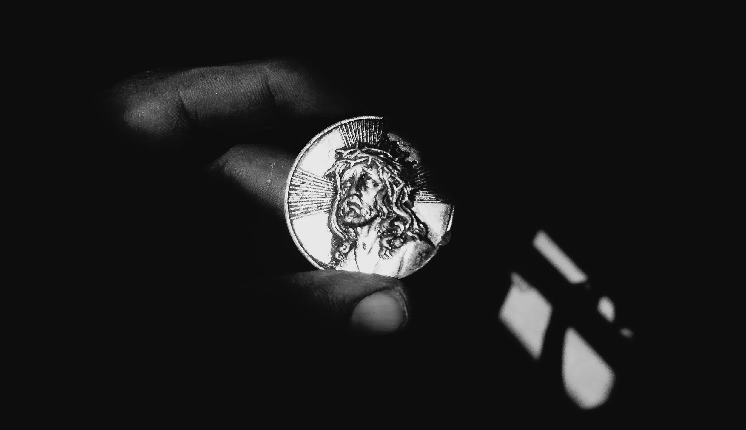Black-and-white photo of fingers holding a medal of Christ crowned with thorns, lit brightly against deep shadow