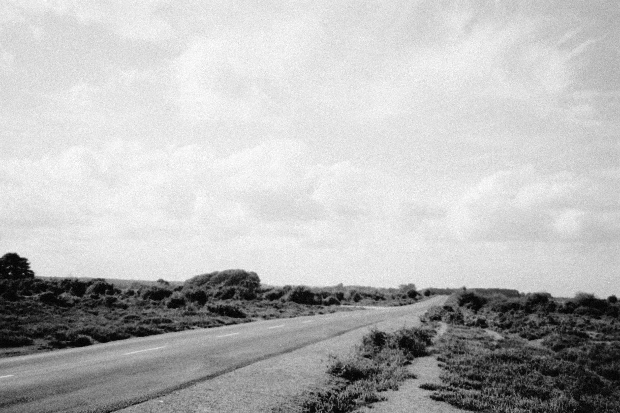 An open road cutting through a low-lying, scrubby countryside beneath a bright, clouded sky in black and white.