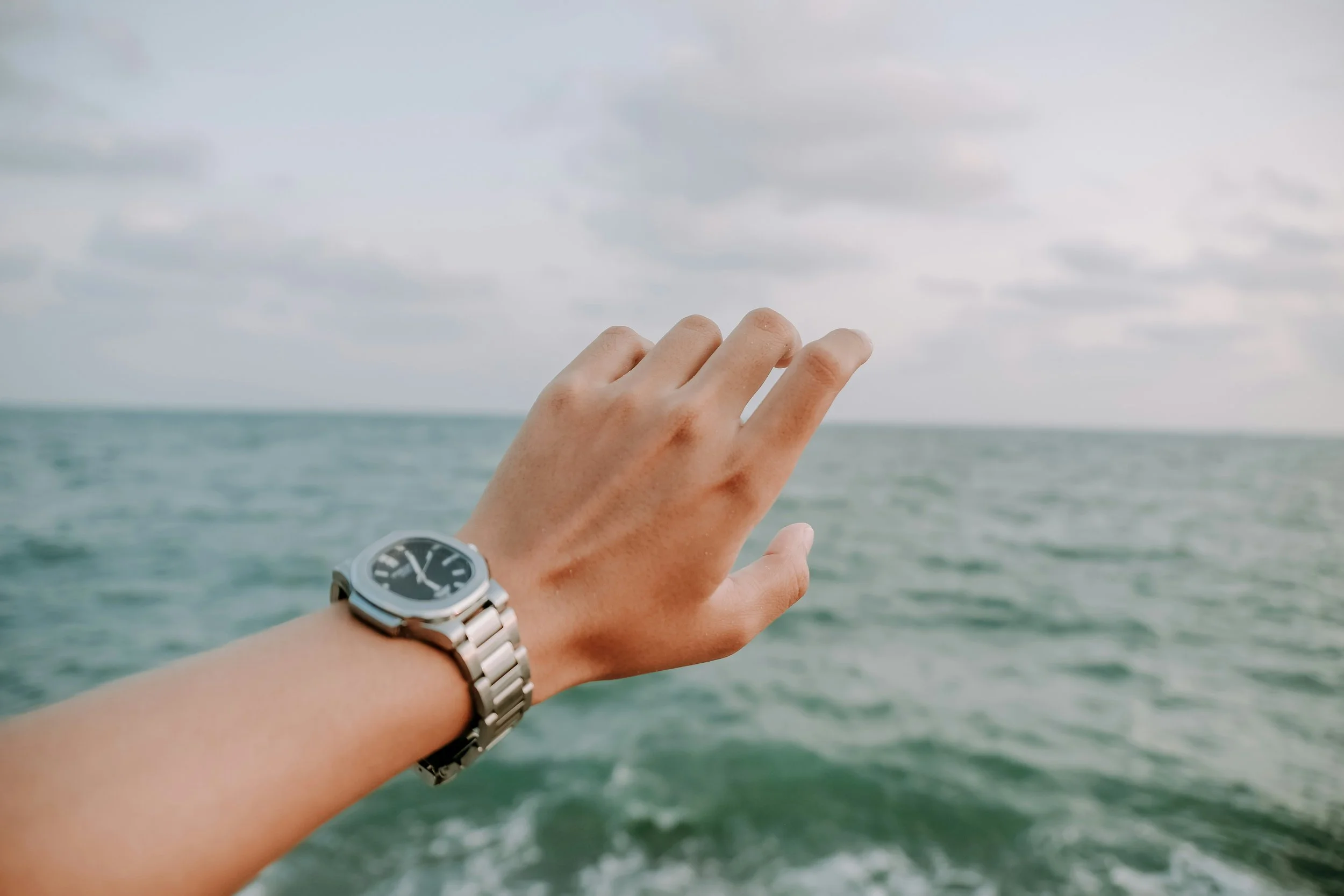 Close-up of an open hand reaching out toward the open ocean with a stainless steel wristwatch on the wrist.