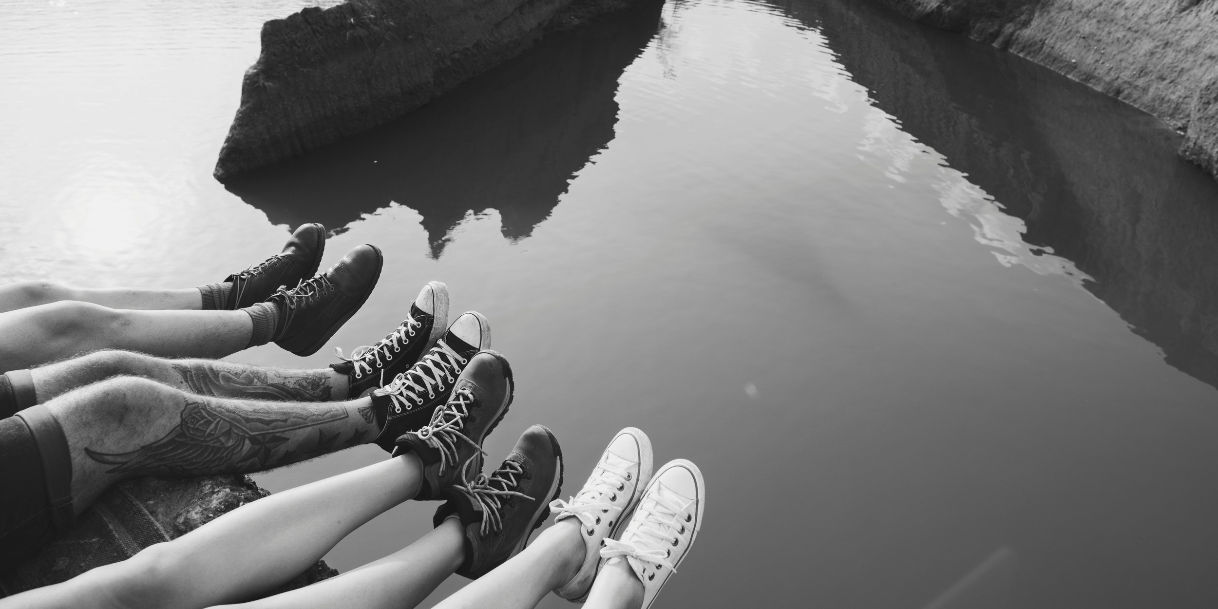 Black and white shot of several distinct pairs of feet dangling side-by-side above a lakeBlack and white photo of friends sitting on a ledge overlooking a lake