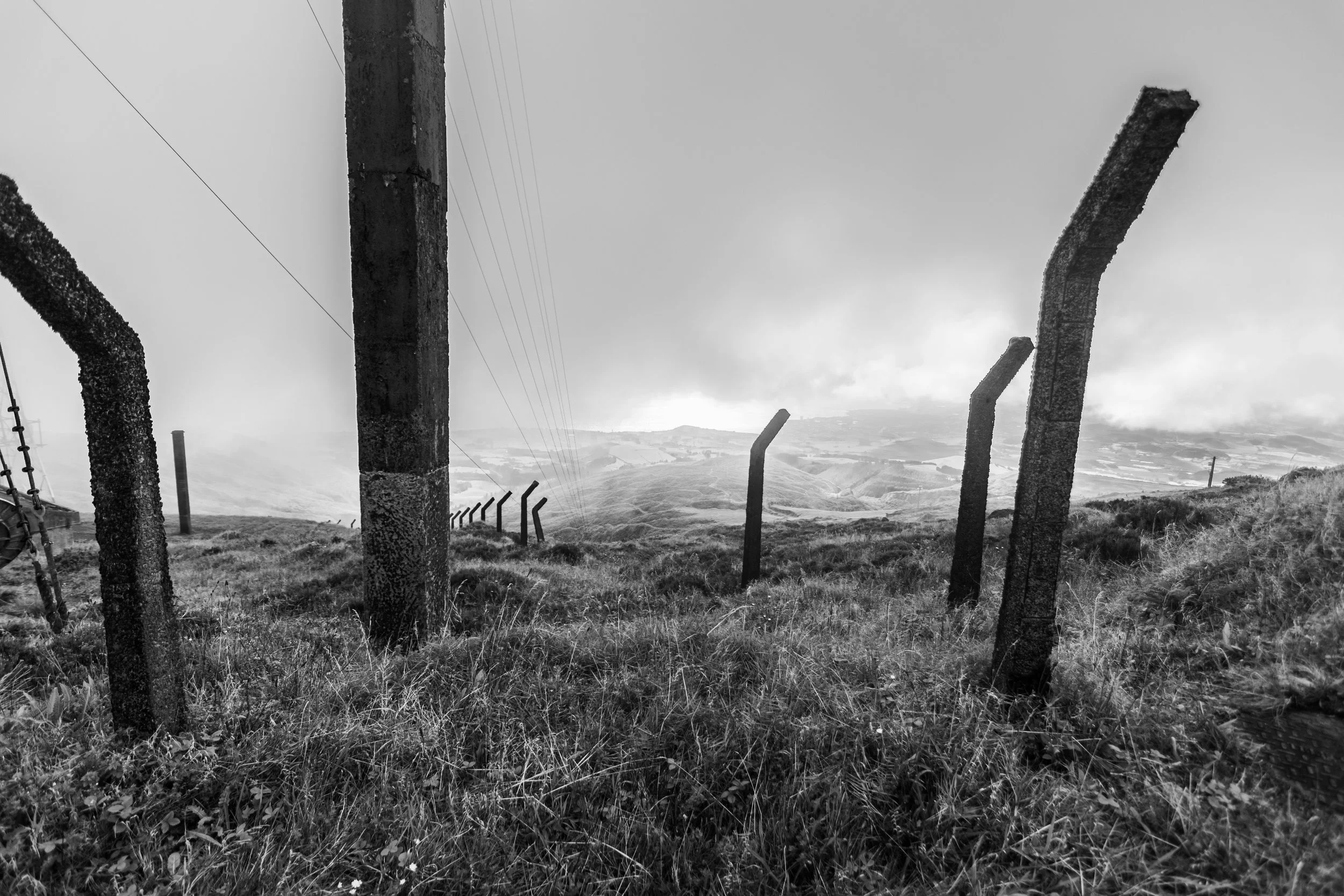 Black-and-white photo of weathered concrete fence posts with bent tops on a misty hillside above a valley