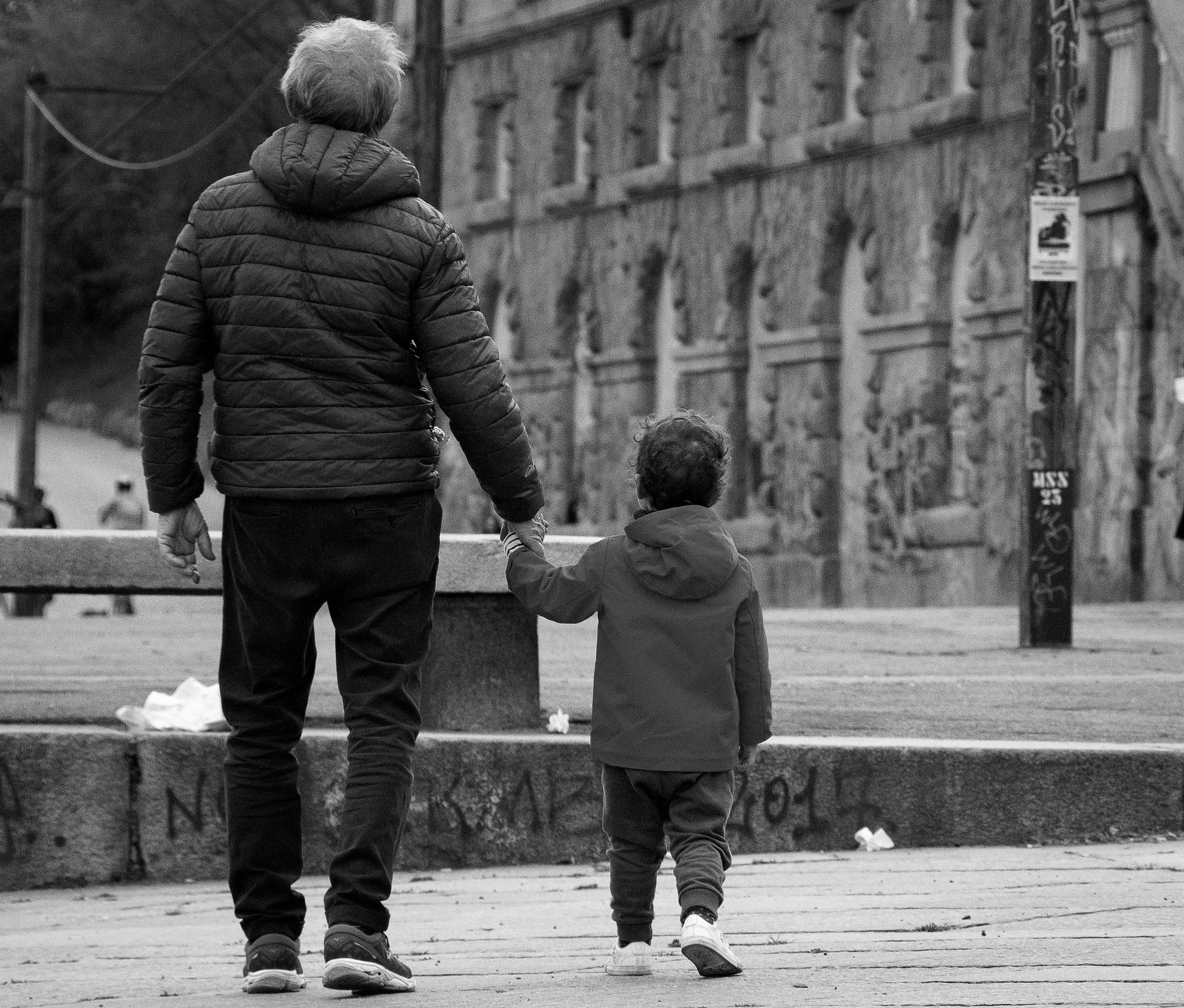 An adult holds a young child’s hand while walking through an urban square in black and white.